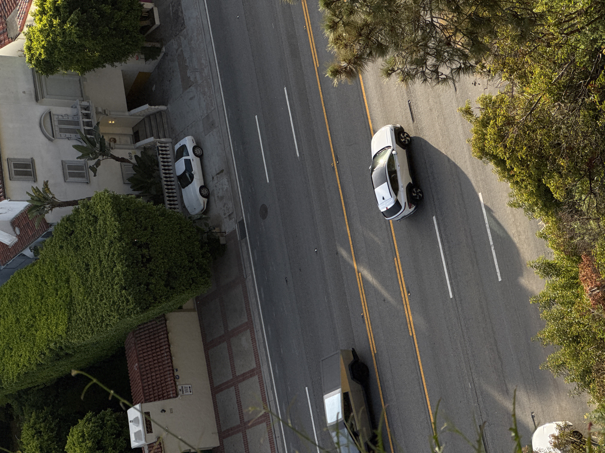 The image shows an aerial view of a street in Santa Monica, California. A white car is parked on the side of the road next to a building, while another white car is driving on the road with a dark-colored vehicle. There are lines on the road, and trees and buildings are visible on either side of the street.