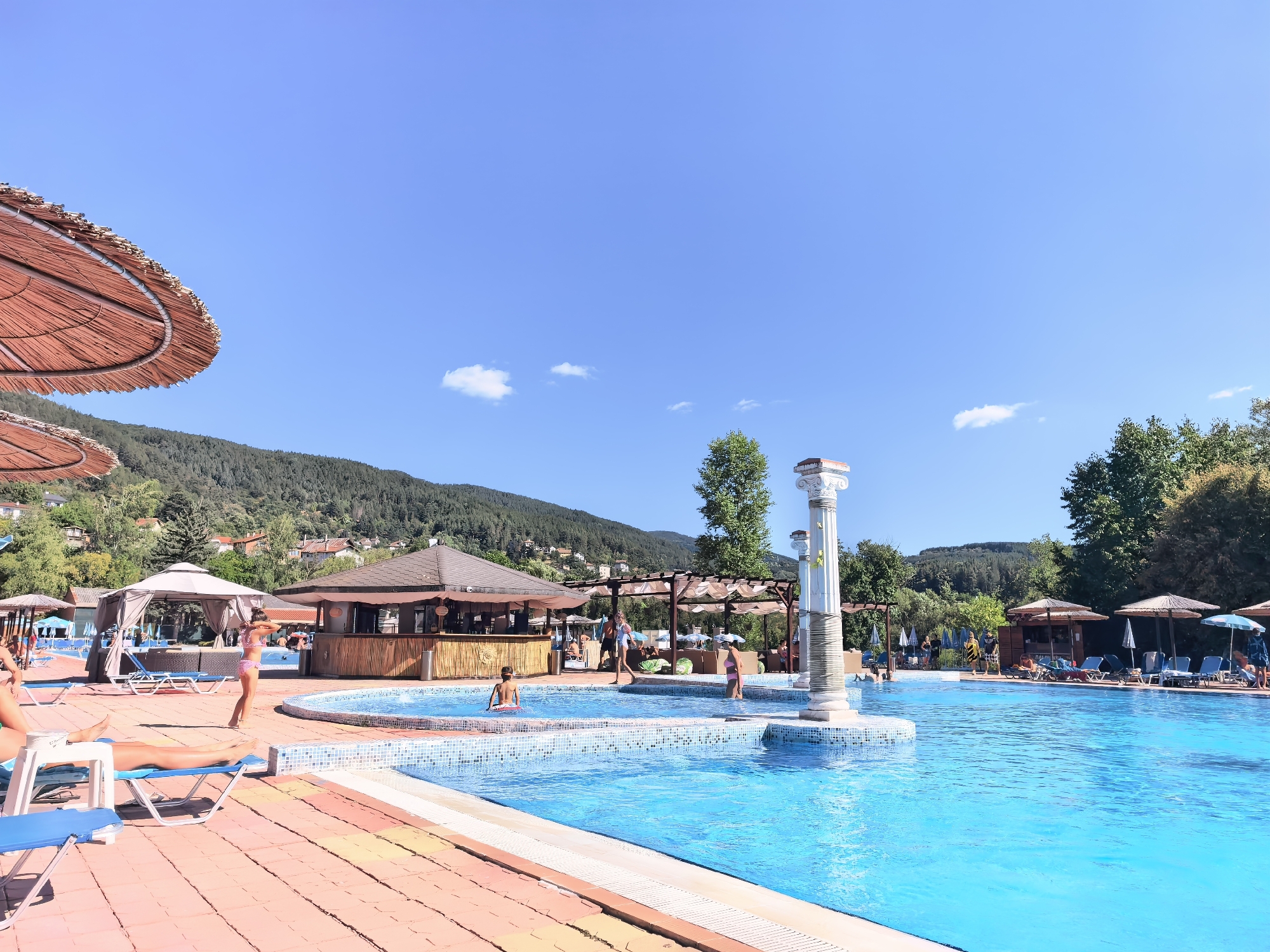 The image depicts a vibrant outdoor swimming pool complex on a sunny day, situated in Rudartsi, Bulgaria. The foreground features a large, bright blue swimming pool with tiled edges. Several people are visible, some lounging on chairs by the pool, while others are in the water. A young girl in a pink bikini stands on the poolside, while a young boy plays in the shallow end of the pool.
In the mid-ground, a bar area with a thatched roof is set up, with people standing at the counter. Several gazebos with white canopies offer shaded seating areas, and lounge chairs with umbrellas are scattered around the pool.
The background is dominated by lush green hillsides covered in trees, with houses nestled among them. The sky is a clear, bright blue with a few wispy clouds. Two large, decorative white columns with ornate capitals are prominent features near the pool, adding a touch of classical architecture to the scene. The overall impression is one of a recreational area designed for relaxation and enjoyment on a warm summer day.