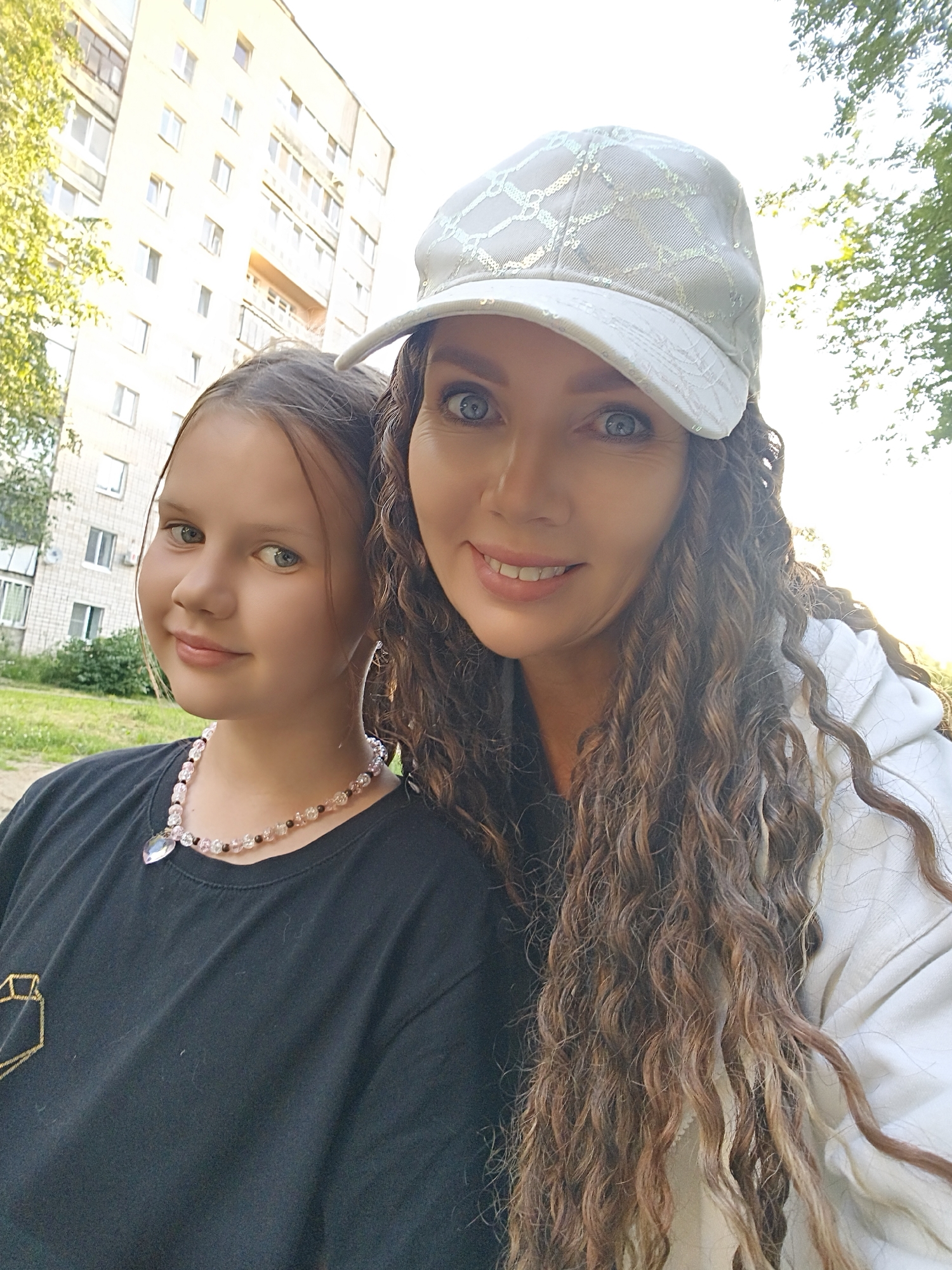 Two people, an adult female and a young female, are posing for a selfie outdoors. The adult female is on the right, wearing a white baseball cap with a textured pattern and a white hoodie. She has long, curly brown hair and bright blue eyes, and is smiling. The young female is on the left, wearing a black t-shirt with a diamond-shaped design and a pink beaded necklace with a heart-shaped pendant. She has brown hair and blue eyes and is also smiling. They are positioned in front of a multi-story apartment building and some trees, with the sky visible in the background. The lighting suggests it is daytime.