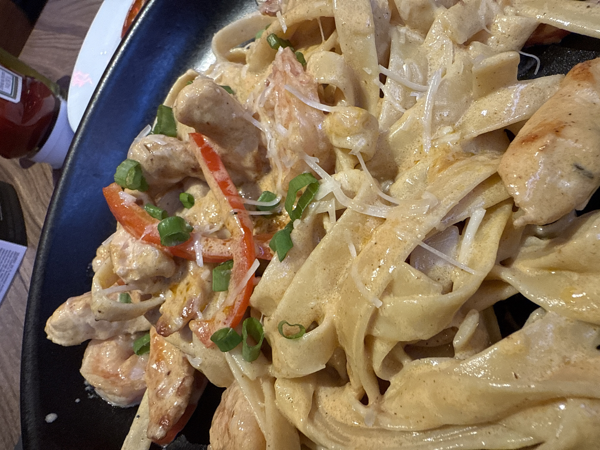 A close-up, high-angle shot captures a dark, possibly black, oval-shaped plate filled with a creamy pasta dish. The main subject is a serving of wide, flat pasta noodles, likely fettuccine or pappardelle, coated in a rich, light orange or beige sauce. Mixed in with the pasta are pieces of what appear to be cooked shrimp and/or chicken, along with sliced red bell peppers and finely chopped green onions (scallions) scattered throughout as a garnish. A generous amount of shredded white cheese, possibly Parmesan, is sprinkled over the top.
The dish appears fresh and warm, with the creamy sauce clinging to the pasta and proteins. The lighting suggests an indoor setting, possibly a restaurant or home dining. In the upper left corner, partially visible, is a red bottle with a white cap, identifiable as a Heinz ketchup bottle, with the word "KETCHUP" visible on its label. A sliver of a white ceramic plate and a wooden table surface are also visible in the background, suggesting a dining environment. No people are visible. The overall impression is an appetizing and hearty meal. The provided location context is San Borja, Peru.