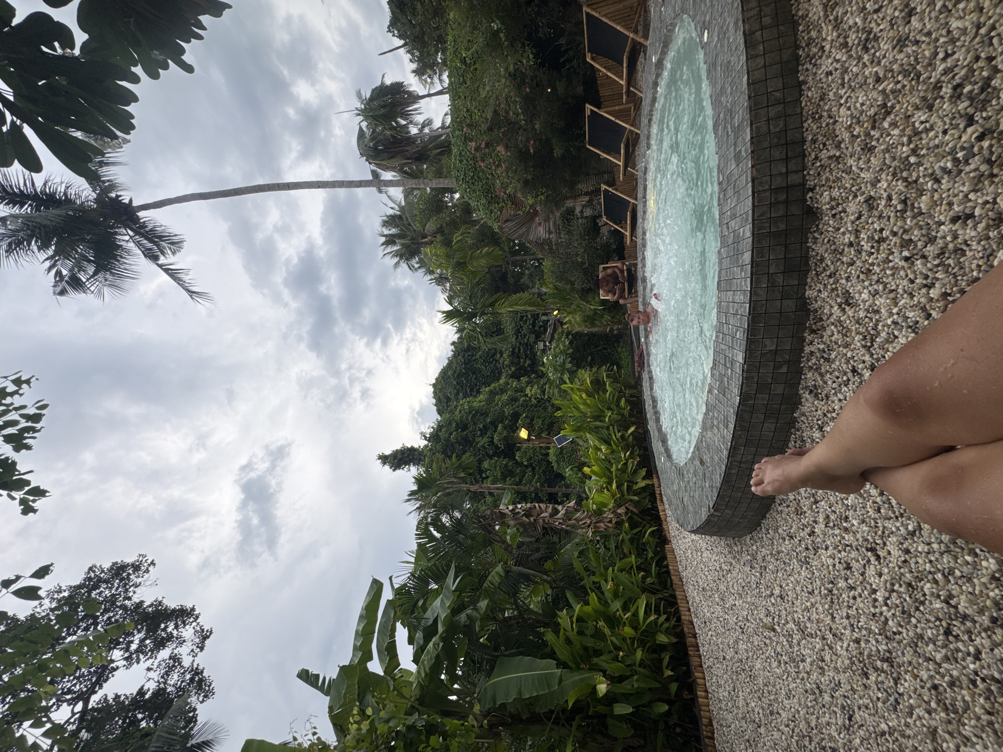 The image depicts a view of a hot tub, likely located outdoors in Baan Sri Thanu, Thailand. A person's legs and foot are visible in the foreground. The hot tub appears to be made of a mosaic tile and filled with water. Two people are visible in the hot tub. The area surrounding the hot tub is covered in small pebbles. Tropical vegetation, including palm trees and banana plants, is visible in the background. The sky is cloudy. Some bamboo furniture is visible next to the hot tub. A small light fixture is also visible amid the foliage in the background.