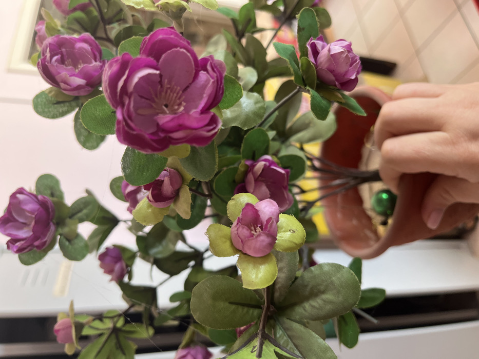 The image displays a close-up, indoor view of an artificial potted plant, assumed to be located in Кез, Russia. A light-skinned human hand is grasping the lower right side of a standard terracotta-colored pot. The pot contains a light-colored granular material, with a small, reflective green spherical object partially embedded within it. The artificial plant features multiple clusters of fabric flowers in shades of purple and magenta, interspersed with green fabric leaves. Fine white strands resembling cobwebs are visible on several leaves and stems. The background is blurred, revealing indistinct light-colored surfaces and a vertical white object on the left, consistent with an interior environment.