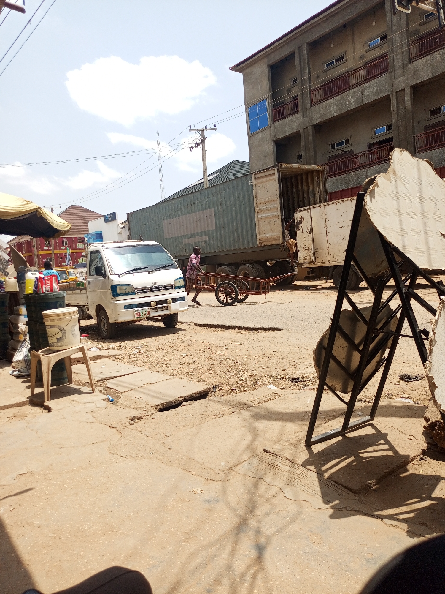 The image captures a street scene in Maiburiji, Nigeria. The foreground is dominated by cracked pavement and a makeshift display stand with large, round objects, possibly stones or concrete forms, leaning against it. A white mini truck with license plate DKA 342 XB is parked on the left side of the street. A man is pulling a two-wheeled cart behind a large shipping container mounted on a truck. Another man is visible inside the container. In the background, there is a multi-story building with rust-colored balconies. Utility poles with wires are visible in the sky, which is partly cloudy. There are goods displayed for sale on the left side of the frame under a yellow awning. A bucket and a small plastic chair are also visible near the storefront. The ground is dusty and there is scattered debris.