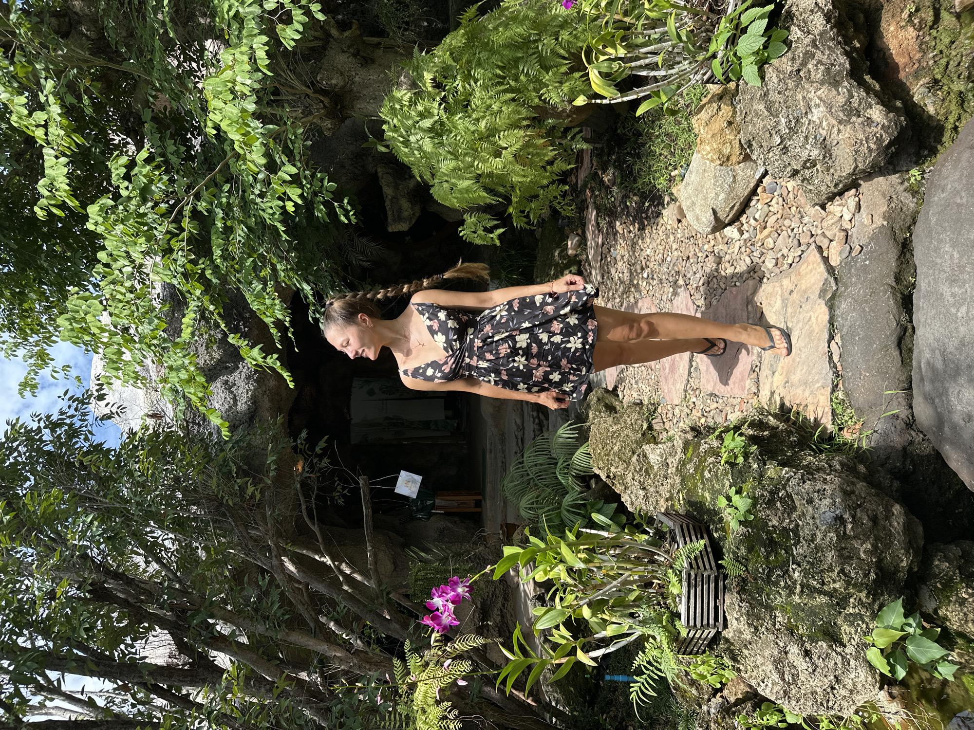 A young Caucasian woman with blonde hair in a braid is walking on a stone path in a lush garden. She is wearing a short, black floral dress and black flip-flops. The garden features large rocks, green plants, ferns, and purple orchids. In the background, there is a cave-like opening with stairs leading up to it. The sky is blue with some clouds. The location appears to be a tropical garden, possibly in Thailand.
