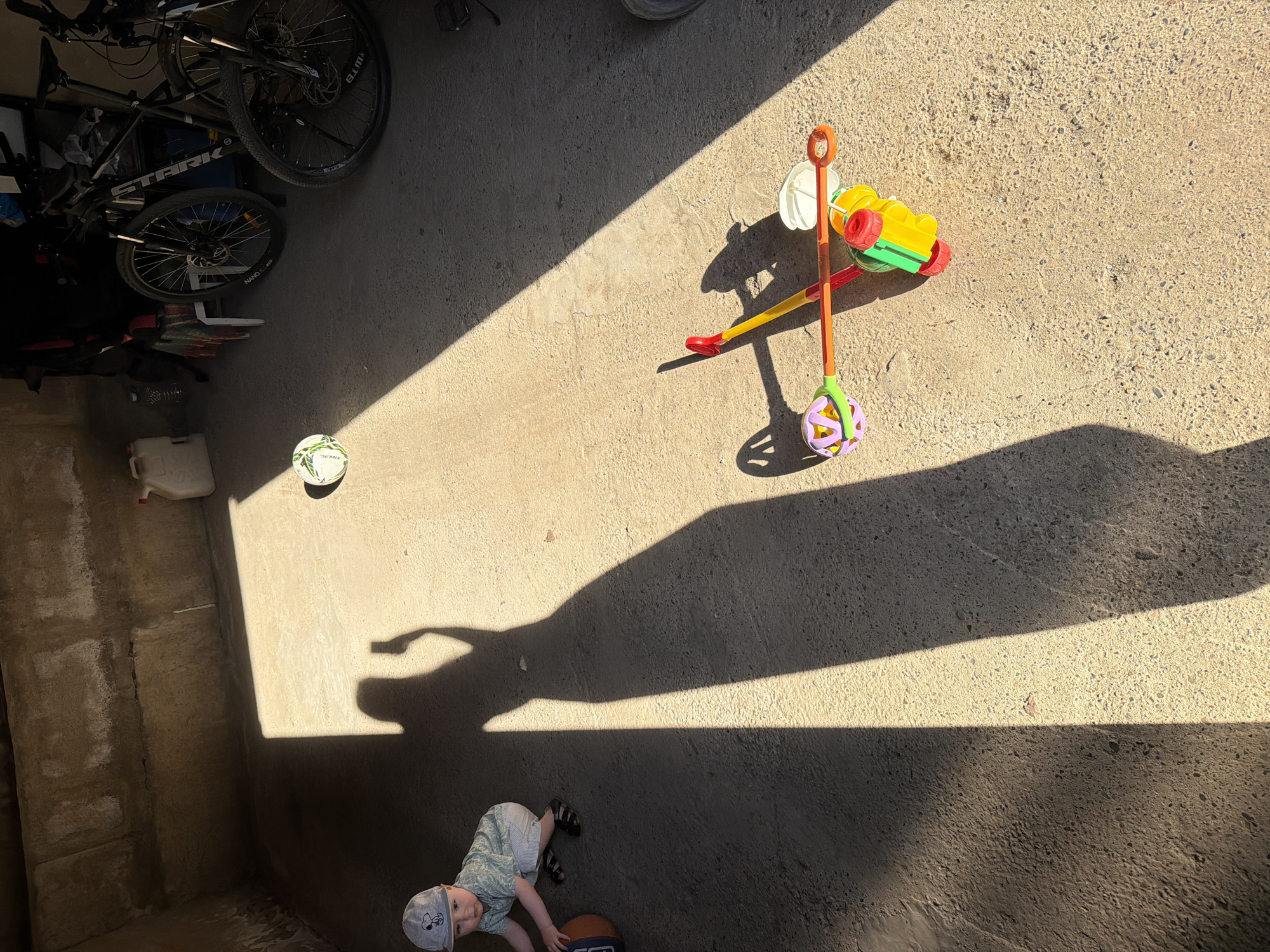 The image shows an outdoor setting, likely a concrete surface such as a patio or garage floor, in the city of Votkinsk, Russia. Natural sunlight casts strong shadows across the area. In the top left are bicycles, a white plastic container and a ball. In the top right is a colorful child's rolling toy. Near the bottom is a toddler playing with a basketball. The shadows suggest it is daytime.