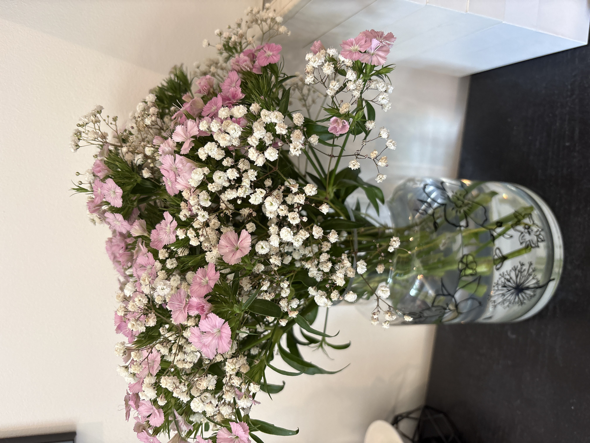 A bouquet of pink dianthus and white baby's breath flowers is arranged in a clear glass vase with black floral illustrations. The vase sits on a black countertop in front of a white wall and a white tiled backsplash. A small portion of a black metal stand and a white object are visible in the lower part of the image. The location is likely indoors, possibly in a home or business in Ipswich, United Kingdom.