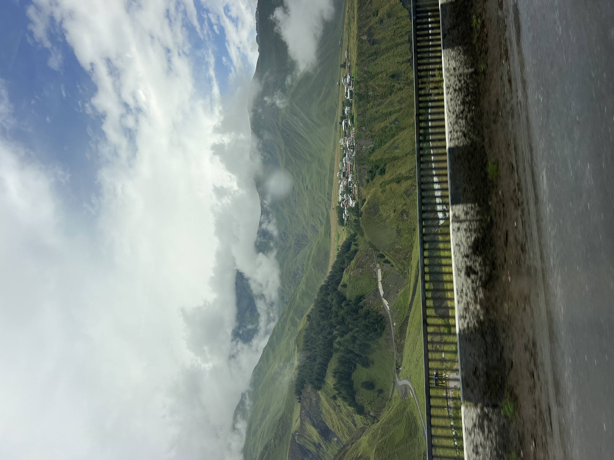 This image depicts a scenic view of the village of Kobi, Georgia, nestled within lush green rolling hills. A paved road is visible in the foreground, separated from the landscape by a railing and a low concrete barrier. The road appears wet, suggesting recent rain. Beyond the road, a verdant valley unfolds, with a winding road cutting through it. In the middle ground, a cluster of houses with red and white roofs forms the village. The background is dominated by majestic mountains, partially shrouded in dramatic, billowing clouds. The sky above is a mix of bright blue patches and dense, overcast clouds, creating a dynamic atmosphere. The overall impression is one of natural beauty and tranquility, with the small village adding a touch of human habitation to the vast mountainous landscape.