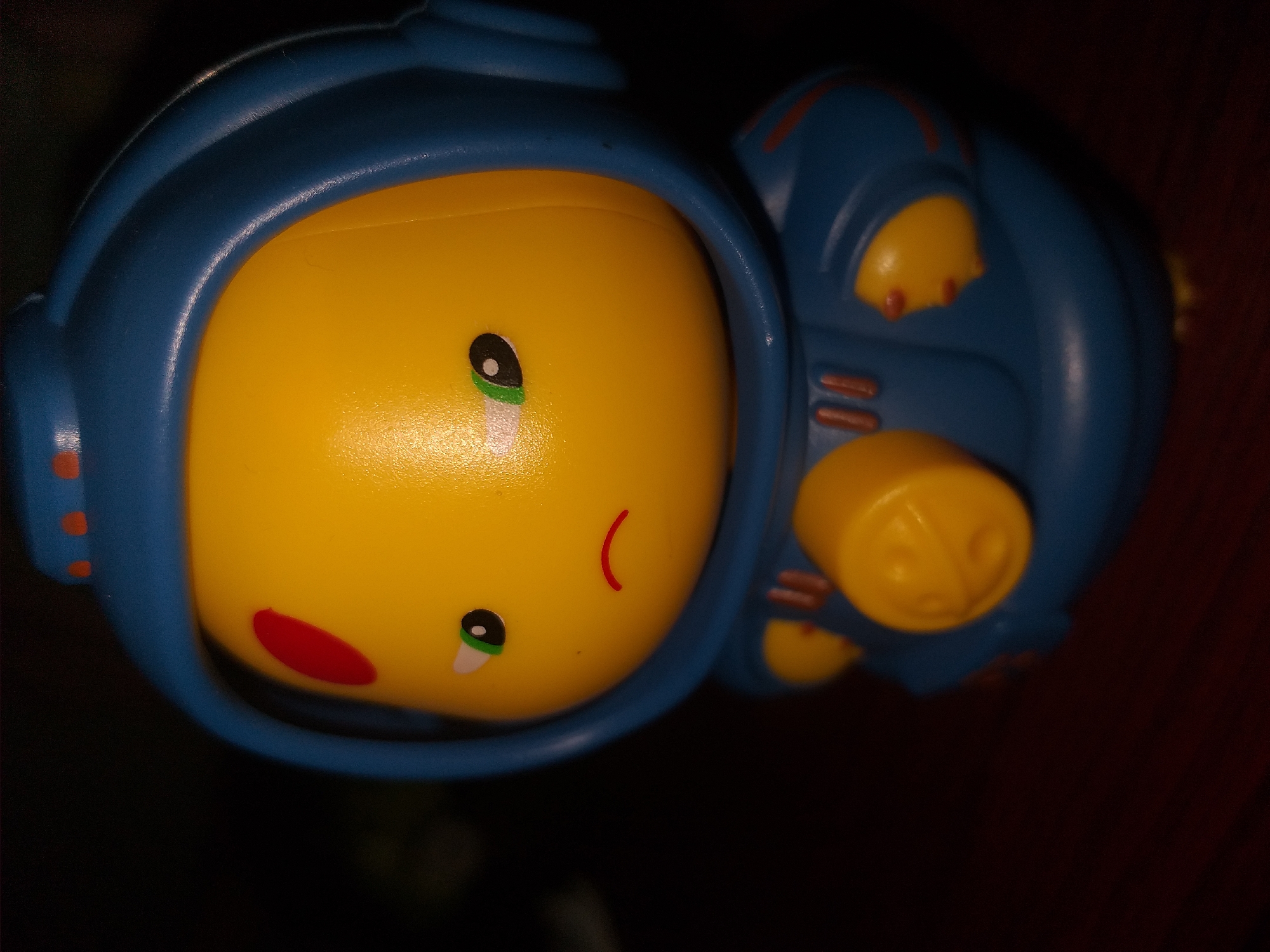 A close-up, vertical, medium shot captures a yellow, plastic toy astronaut with a blue helmet and suit. The toy's face is round and yellow, with one green and black eye on the left, a black pupil on the right, and a small red curved line for a mouth. A red circular marking is present on the left side of its forehead. The blue suit has orange accents and a circular yellow button-like feature on the chest with a molded design. The toy is positioned slightly to the right of the frame, with its body angled towards the viewer. The background is dark and out of focus, suggesting a dimly lit indoor environment. The surface on which the toy rests appears to be a dark brown, wooden table. The lighting creates shadows, highlighting the texture of the plastic. This image is not located in Air Jamban, Indonesia, as there is no discernible urban environment or specific geographical markers to suggest such a location.