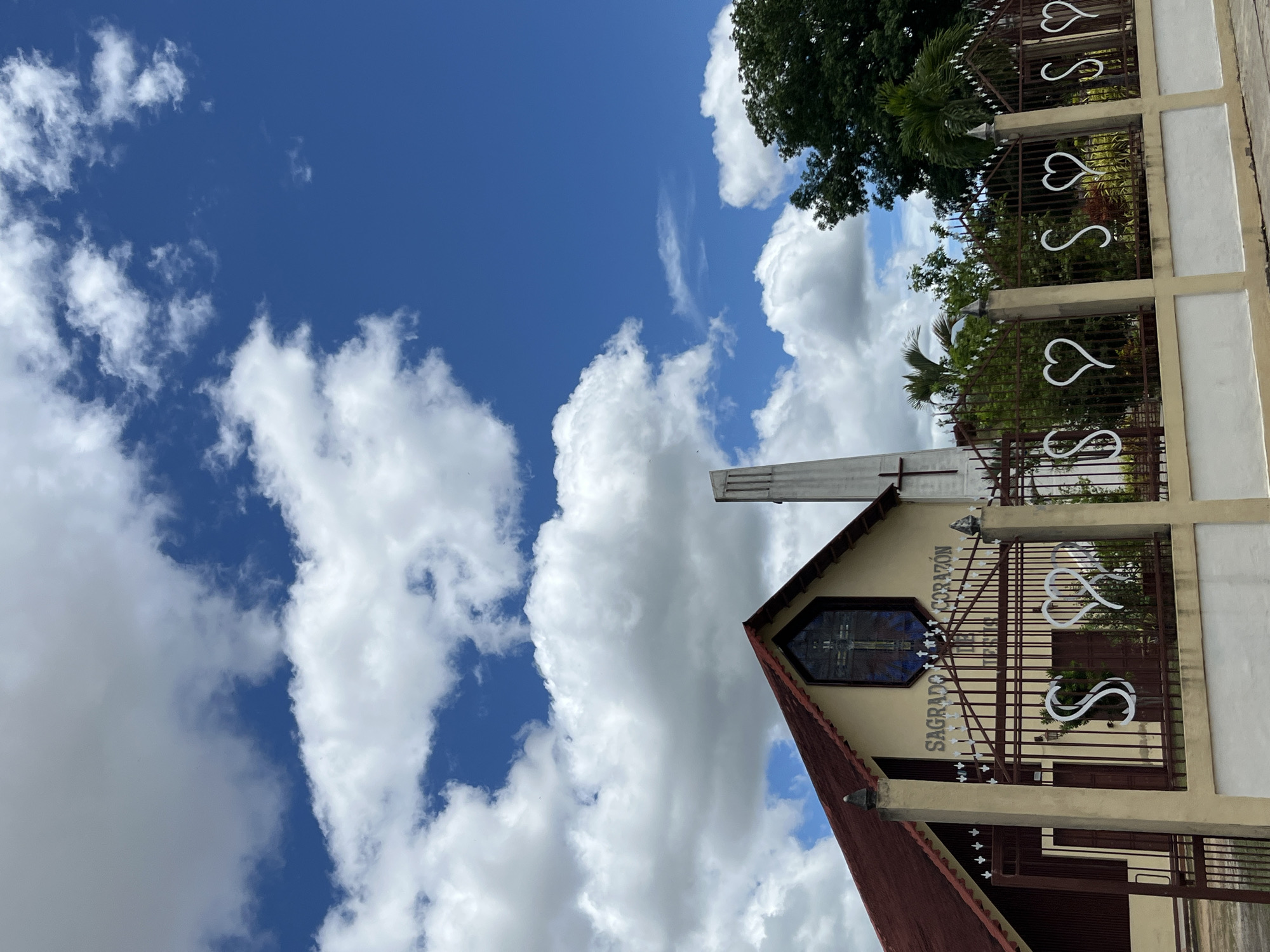 The image captures the facade of a church in Tinaquillo, Venezuela, under a bright, partly cloudy sky. The church, painted in shades of yellow and beige, features a prominent red-tiled roof and a stained-glass window depicting a cross. A tall, slender bell tower stands to the right of the main structure. A decorative iron fence, adorned with stylized heart shapes, runs across the foreground, separating the church from the viewer. Lush green trees and tropical foliage are visible behind the fence and surrounding the church. The sky is a vibrant blue, dotted with fluffy white clouds, suggesting a clear and sunny day. The overall scene conveys a sense of peace and religious devotion within a tropical setting.