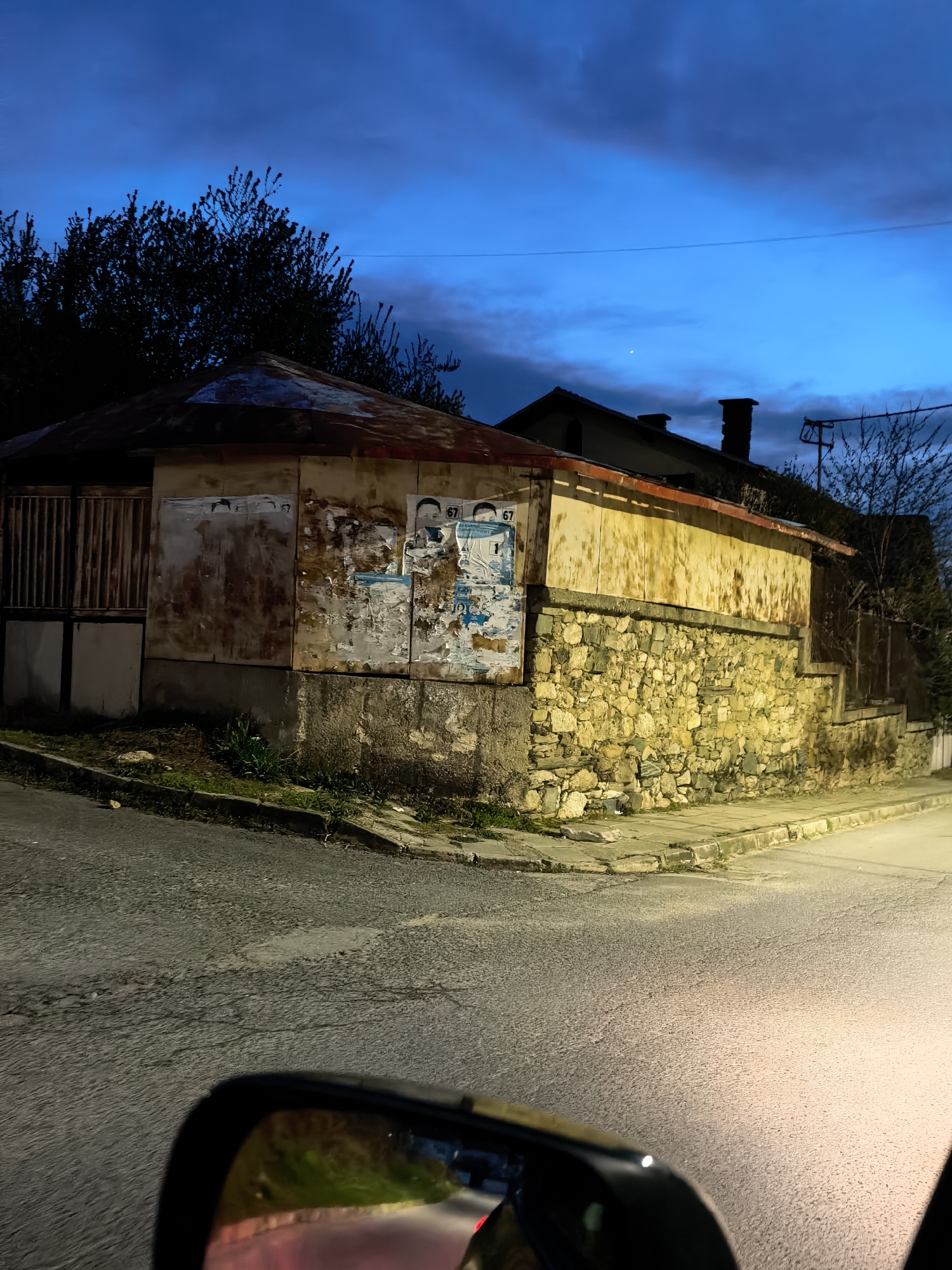 The image presents an evening street scene in an unspecified urban area. The foreground includes a portion of a vehicle's side mirror, reflecting a blurred image of red and green lights, suggesting a road or possible traffic. The street itself is dimly lit, showing the textured asphalt surface and its intersection with a sidewalk.
In the midground, a low, stone building stands on the corner. The building's lower section is constructed from stacked, rough-hewn stones, while the upper portion appears to be made of concrete panels or cinder blocks, some covered with posters and peeling surfaces. The building is topped with a rust-colored, corrugated roof. Adjacent to the building is a wooden fence or gate, which appears to be weathered.
The background features silhouetted trees against a blue, twilight sky, hinting at a time shortly after sunset. A second building, possibly residential, is partially visible behind the stone building, with a chimney extending above its roofline. Utility poles and wires are also seen against the sky, indicating a settled, though possibly aged, urban landscape. The overall impression is one of a quiet, possibly overlooked part of a town at dusk.