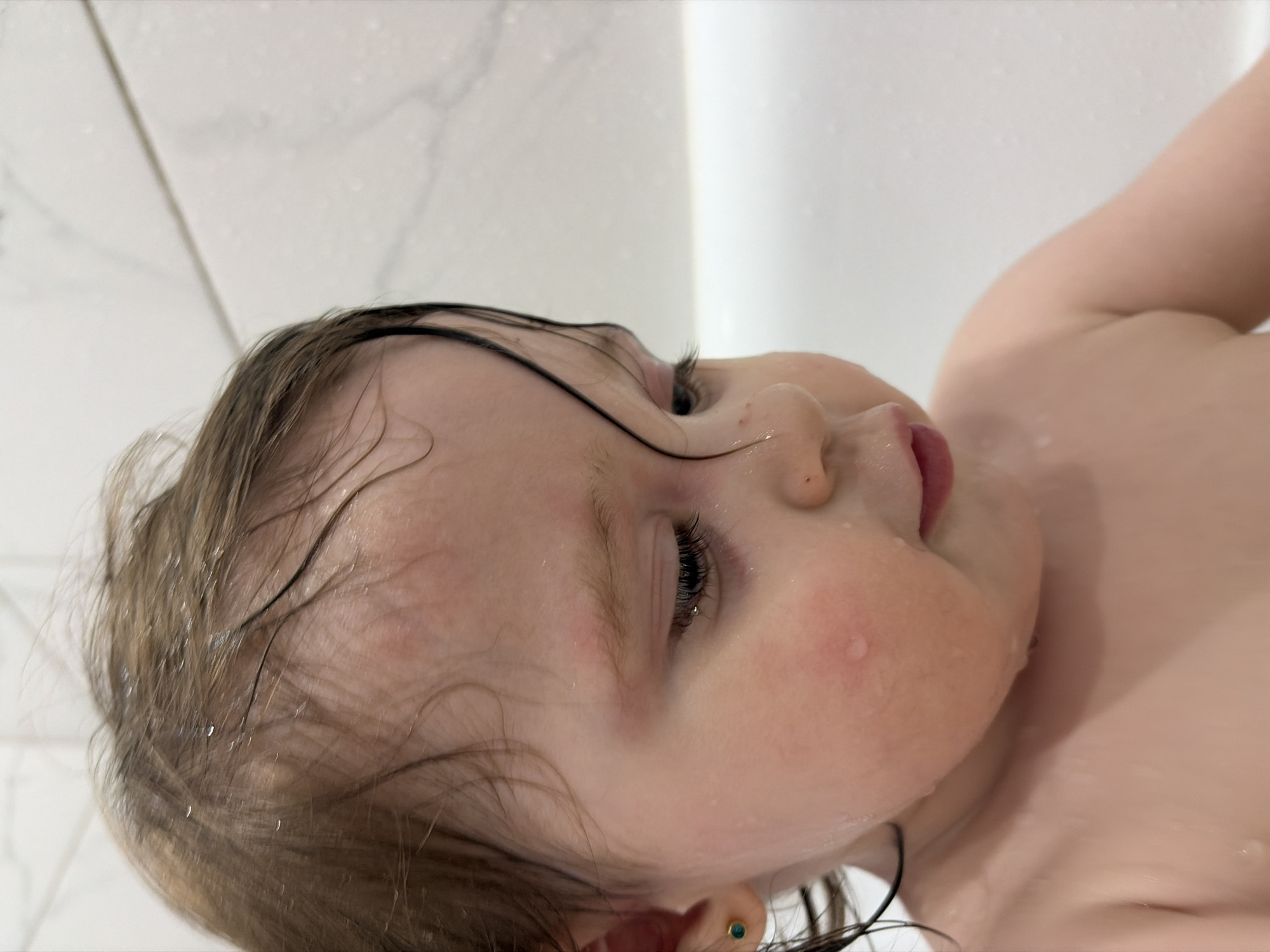 A young child, possibly a toddler, is depicted in a wet environment, likely a bathtub or shower. The child has light skin, wet hair, and is angled towards the viewer, showing their face, including eyes, nose, and partially open mouth. There are droplets of water on their face and hair. A small, possibly decorative earring is visible in the ear. The background appears to be a white tiled wall. No information about the location in the city of Chaikovsky, Russia is available in the provided image.