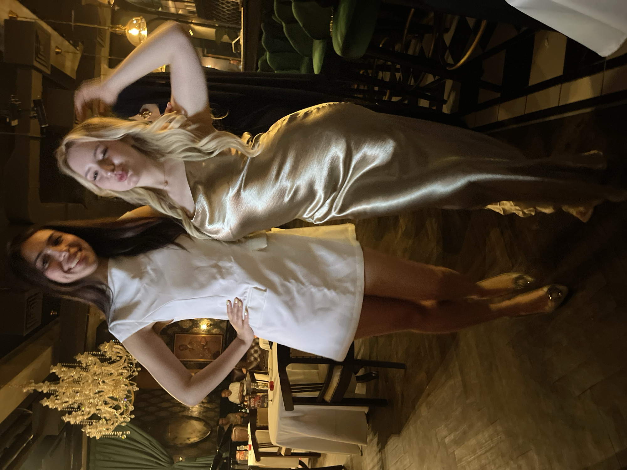 Two young women are posing for a photo in what appears to be a dimly lit, upscale restaurant or lounge with a chandelier. The woman on the left is wearing a short white dress and high heels. She is smiling and has her left hand on her hip. The woman on the right is wearing a long, shimmering gold dress and has her hair down. She is puckering her lips and has her right hand raised, possibly touching her hair. In the background, there are tables with white tablecloths, chairs, and a bar area with green velvet stools. The flooring is a dark wood parquet. The overall ambiance suggests a social gathering or night out. The location is identified as South Jakarta, Indonesia.