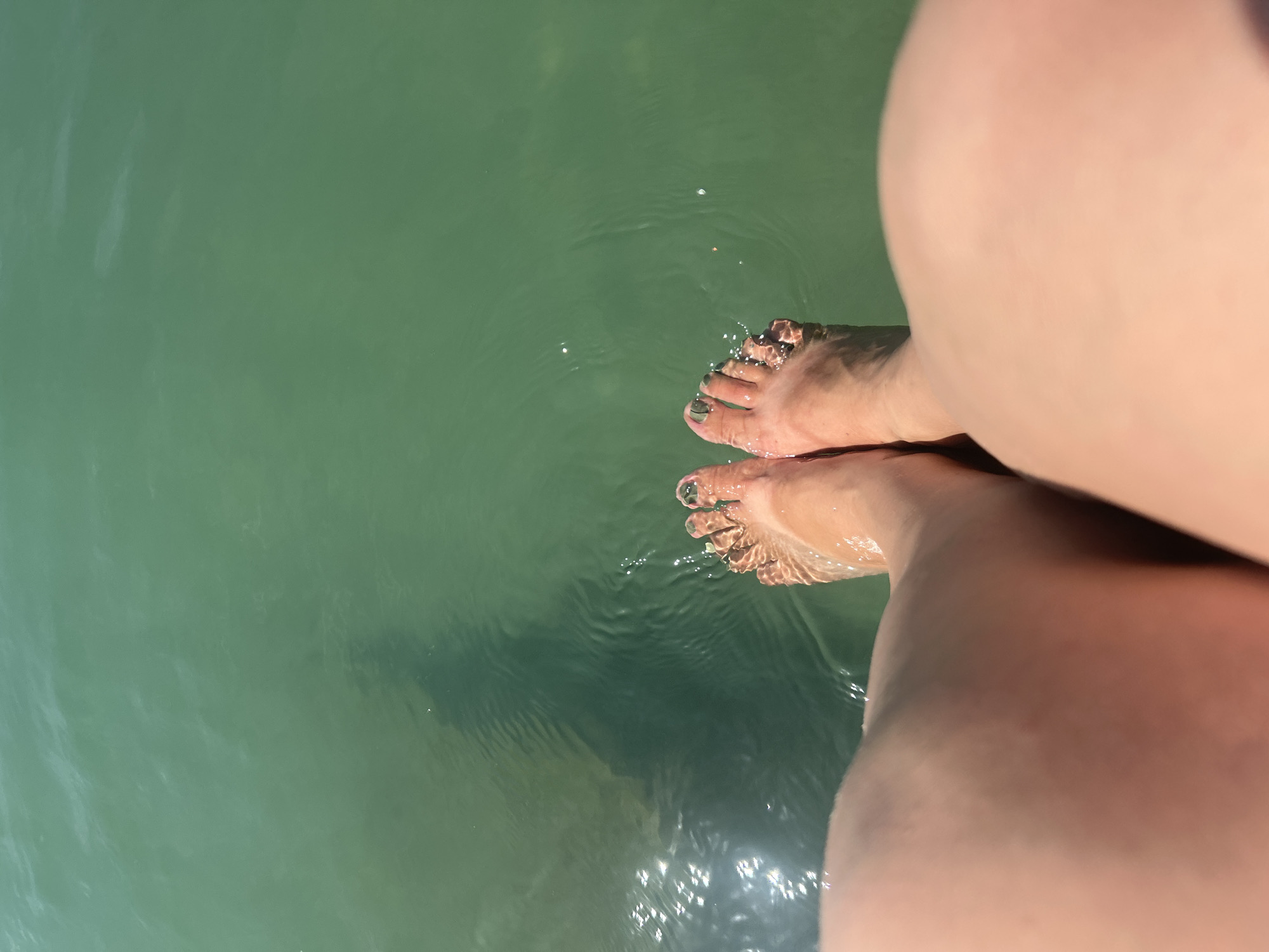 In Katoro, Croatia, a close-up aerial shot shows a person's legs and feet submerged in shallow, clear green water. The water's surface is rippled, with sunlight reflecting off it. A faint shadow of a person is visible beneath the water's surface, extending from the person's feet. The toes are painted with dark green nail polish. The scene is devoid of any other subjects or objects.