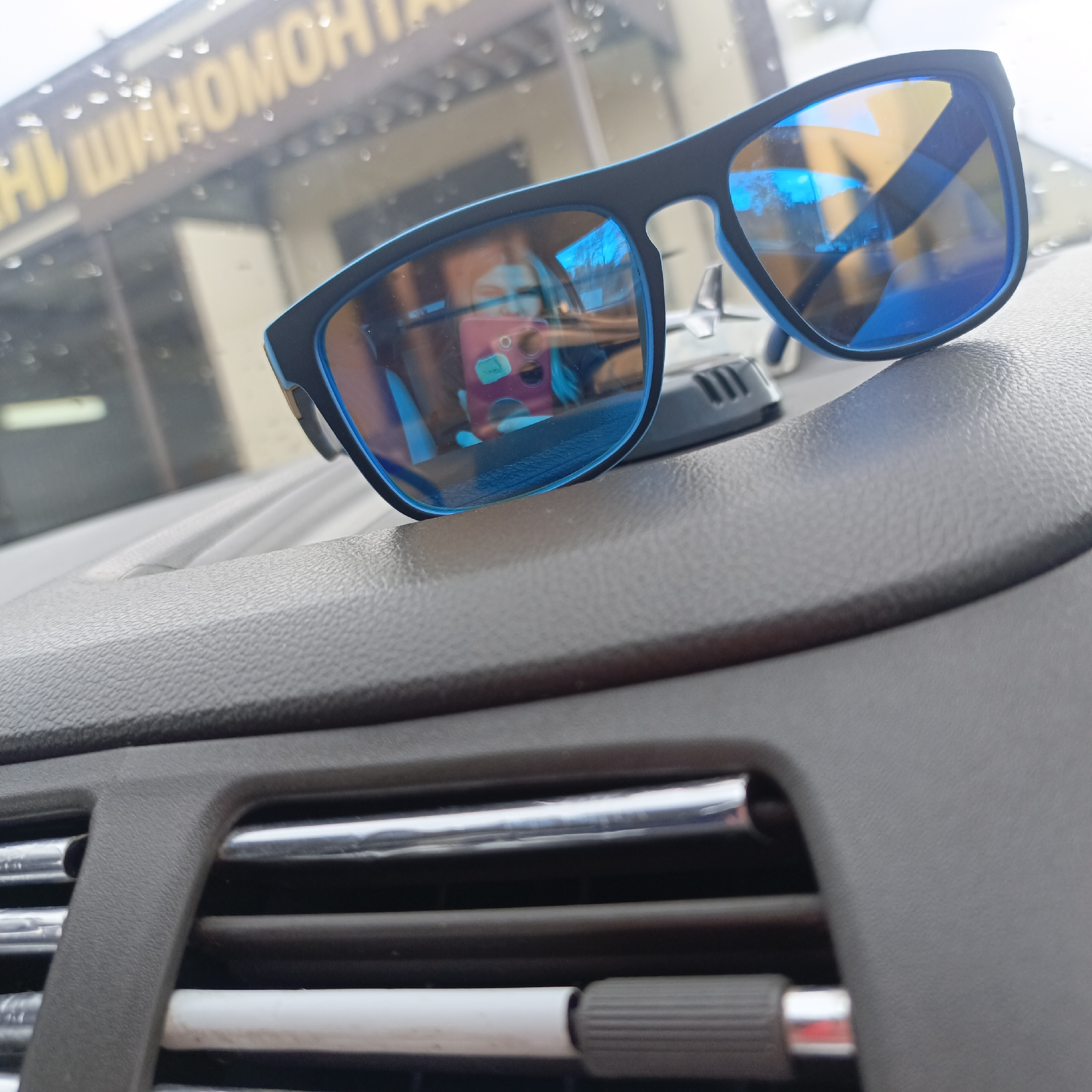 The image shows a pair of sunglasses with blue lenses and black frames resting on the dashboard of a car. The air conditioning vents of the car are visible in the foreground. The reflection in the sunglasses shows a person holding a phone, presumably taking the photo. In the background, a building is visible with signage in a Cyrillic alphabet. Based on the script, the location is in a city of Unknown, Unknown. The weather appears to be overcast or rainy, indicated by droplets on the windshield.