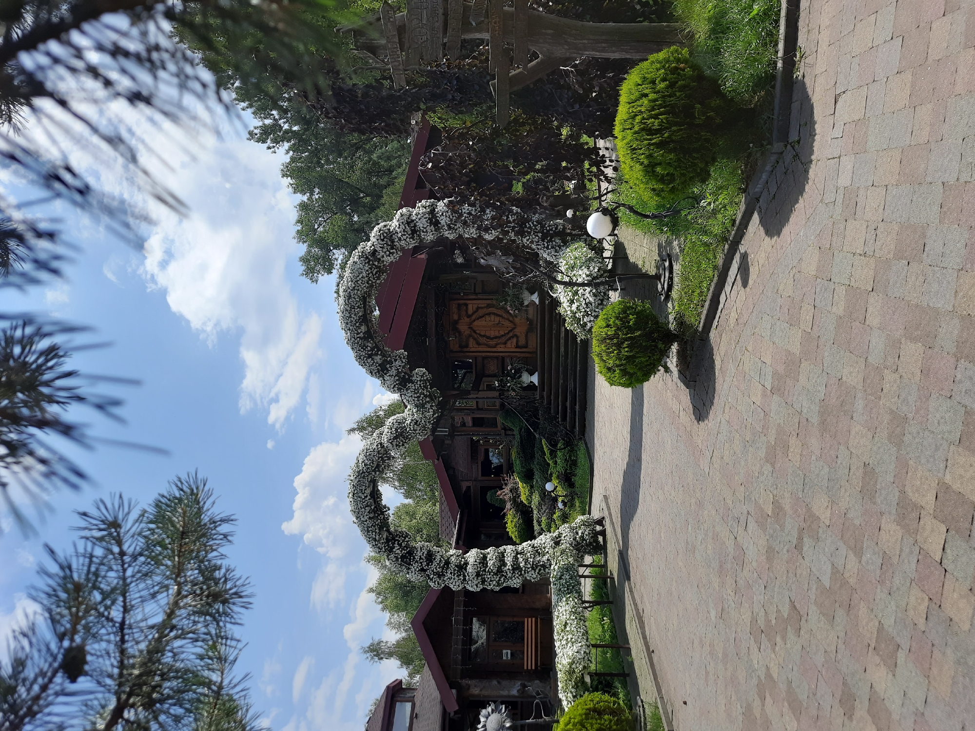The image shows an outdoor scene at the entrance of a restaurant in Drohobych, Ukraine. A large, heart-shaped arch decorated with white flowers is the focal point of the scene, leading towards the entrance of a wooden building. The building appears to be a traditional Ukrainian restaurant, with a wooden facade and a dark red roof. A set of stairs leads up to the main entrance, which is adorned with a large, intricately carved wooden door. Surrounding the entrance are manicured bushes, green lawns, and decorative white spheres on stands. In the foreground, a paved walkway made of multi-colored bricks leads to the arch. Trees and other greenery form the backdrop of the scene, with a blue sky and scattered clouds above. On the right side of the image, a wooden signpost with multiple directional signs is visible. The overall impression is one of a festive and welcoming atmosphere, likely for a special event or celebration.