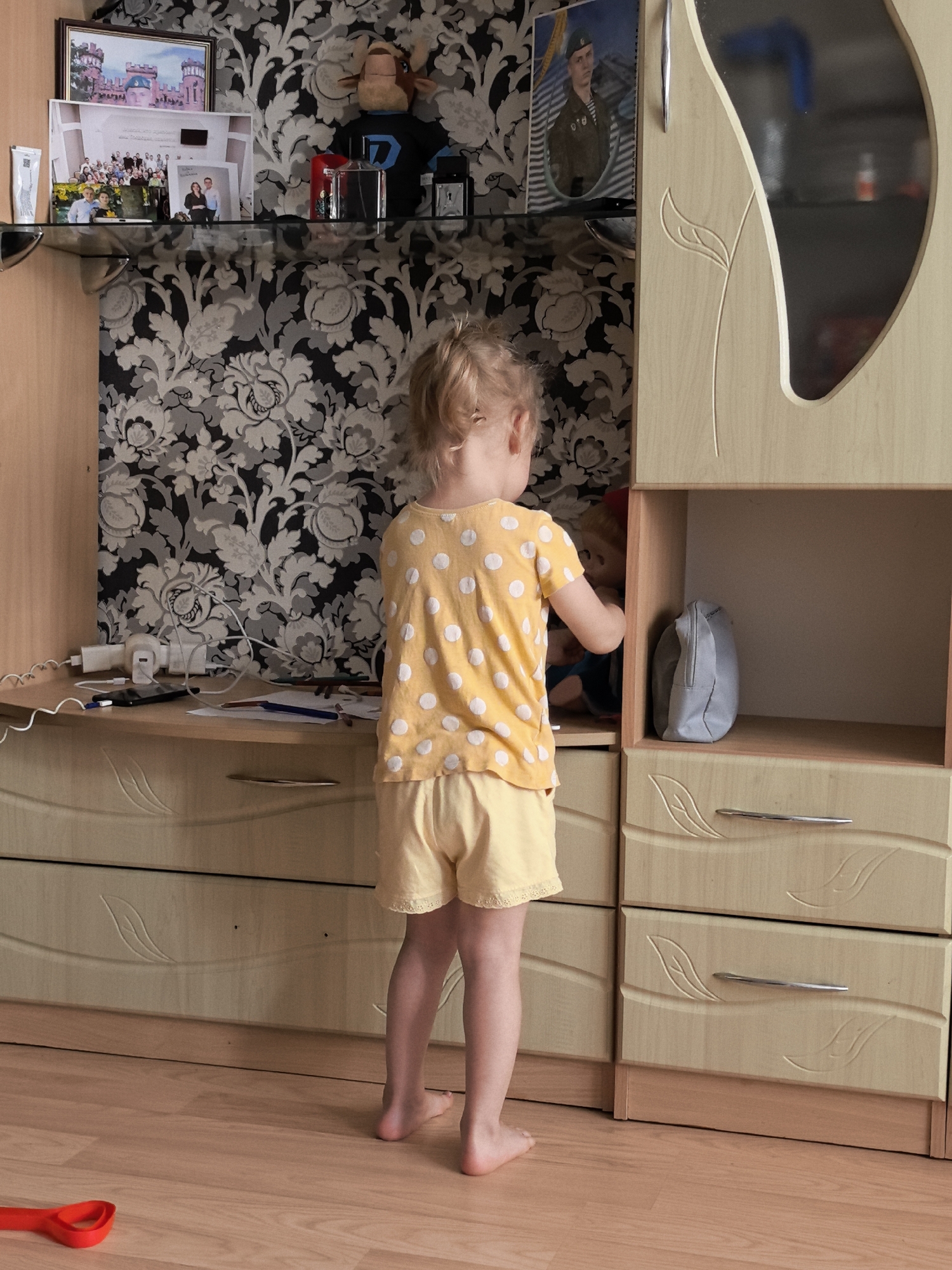 A young child stands facing a light-colored wooden cabinet with carved leaf designs on its drawers and doors. The child, wearing a yellow polka-dot t-shirt and yellow shorts, reaches into an open shelf of the cabinet, where a gray pouch rests. The cabinet is situated against a wall covered in black and white floral wallpaper. A desk with papers, charging cables, and pens is visible to the left of the cabinet. Above the desk, shelves hold framed photos, a stuffed animal, and decorative items. The floor is a light wood laminate. The overall scene appears to be an indoor domestic setting, possibly a living room or bedroom.