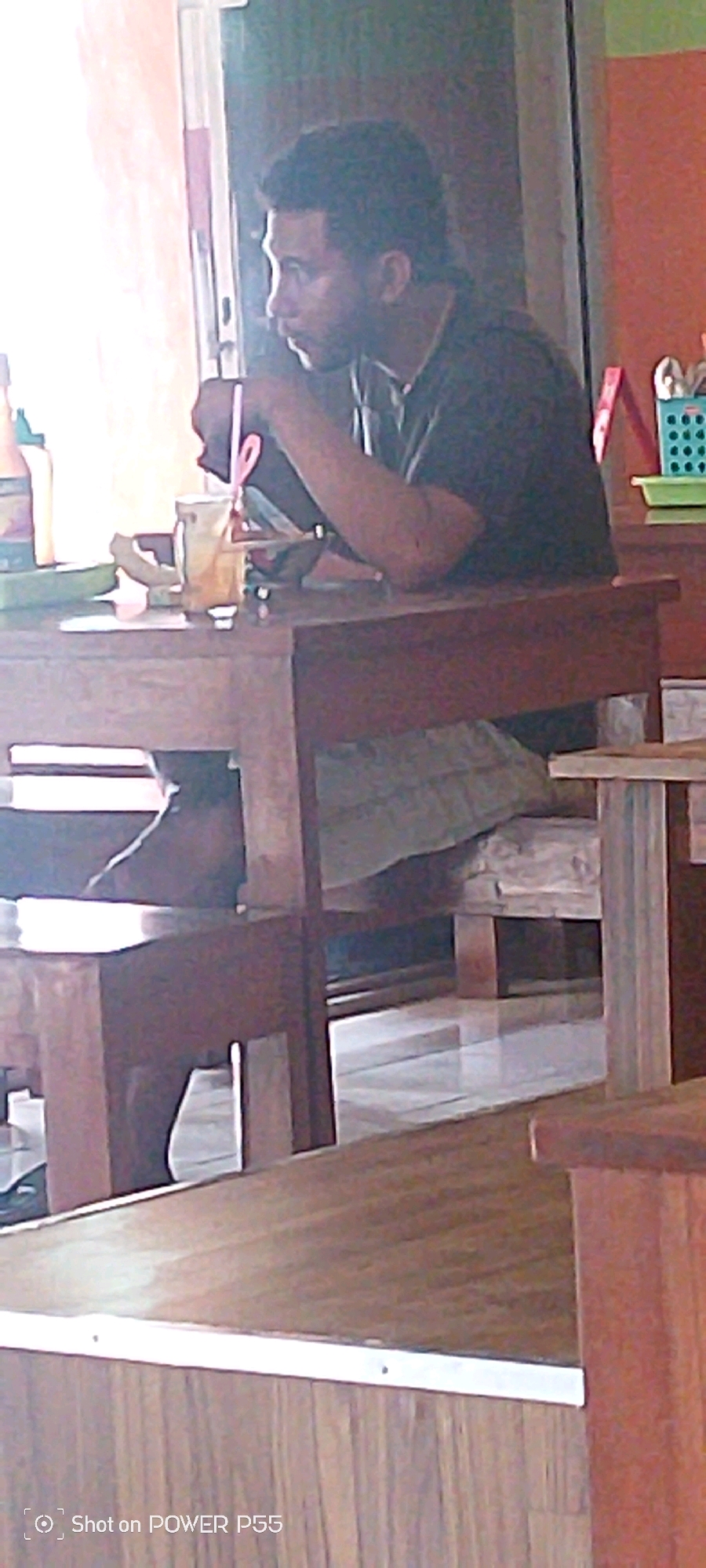 In Secang, Indonesia, a person sits at a wooden table with a drink. The person is wearing a dark t-shirt and appears to be looking down at the table. A glass with a straw is visible on the table, along with some other items that are not clearly identifiable. The environment appears to be indoors, with a bright light source coming from the left side of the image. The overall scene suggests a casual setting, possibly a cafe or restaurant. The image is low resolution and has a slightly blurred quality.