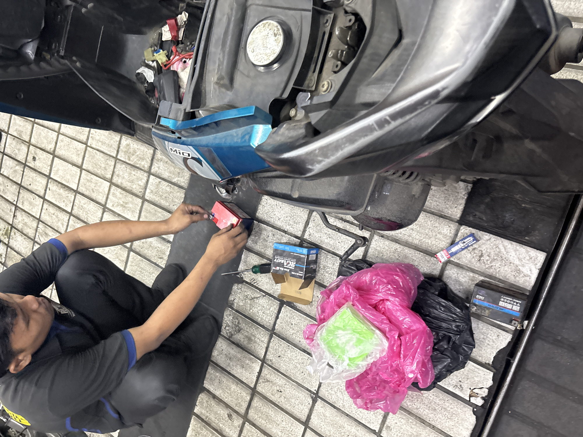 In a workshop setting in North Jakarta, Indonesia, a male mechanic wearing a black and blue uniform is kneeling on a tiled floor next to a disassembled scooter. The scooter's seat is open, revealing its internal components. The mechanic is holding a red battery and a screwdriver, suggesting he is in the process of replacing or working on the scooter's battery. Various items are scattered around the workspace, including a blue battery box labeled "RK.BATT," a green object in a plastic bag, pink and black plastic bags, and a spark plug. The floor is made of beige and white square tiles, and the overall impression is one of a busy automotive repair environment.