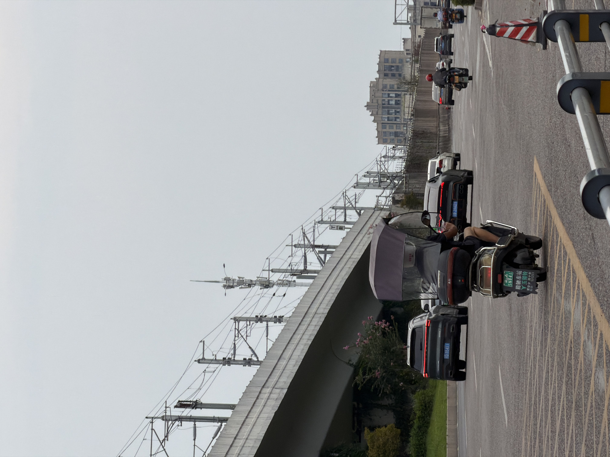 The image shows a street scene in the city of Jiangjin, China, under an overcast sky. A multi-lane road is visible with several vehicles in motion. In the foreground, a person is riding a three-wheeled electric scooter with a canopy. Behind it, a dark-colored SUV with a red taillight bar is driving away. Further down the road, other cars and motorcycles are visible, including a motorcycle with a rider wearing a red helmet. A concrete overpass, likely for a railway or elevated road, runs across the upper left portion of the frame, supported by numerous poles and wires. In the background, a large, old, multi-story building with many windows stands against the sky. The road surface has yellow and white lane markings.