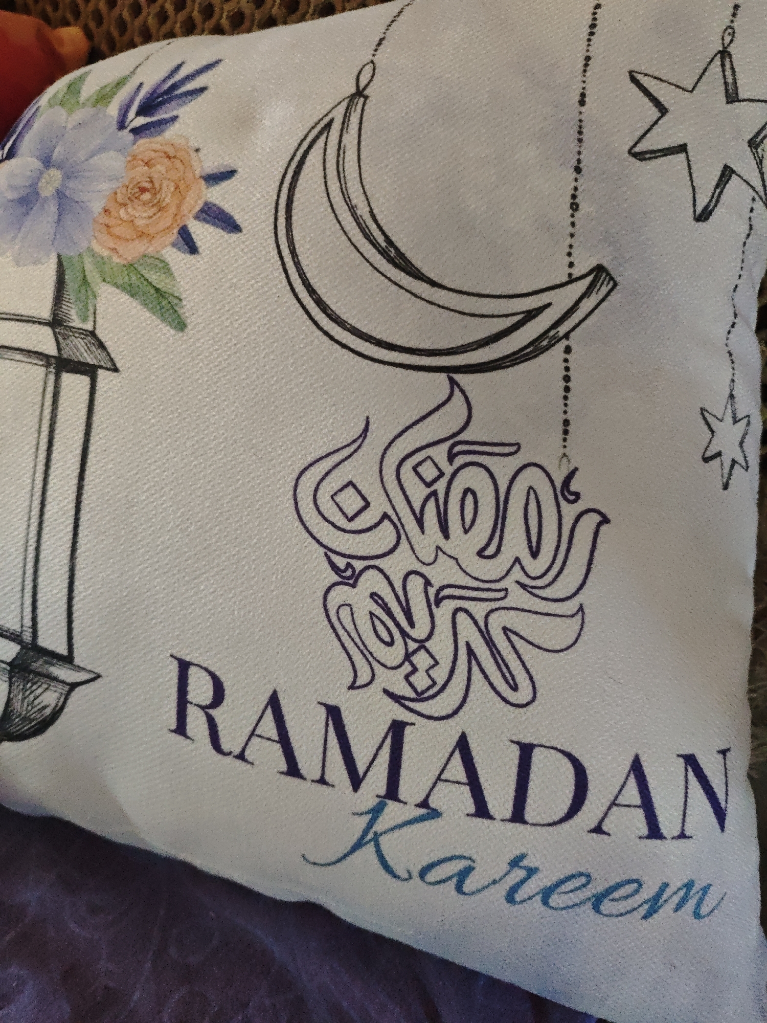 A close-up of a white cushion with Ramadan-themed decorations. The cushion features a drawing of a crescent moon in black outline, an intricate purple Arabic calligraphic design, and the words "RAMADAN Kareem" printed beneath it. The word "RAMADAN" is in dark purple, while "Kareem" is in blue. Additionally, there is a drawing of a lantern with floral elements in the corner. A string of decorative beads hangs from the crescent moon, and a star is depicted near the top right corner. The background is blurred. There is no information about the location in the city of Боровково, Russia.