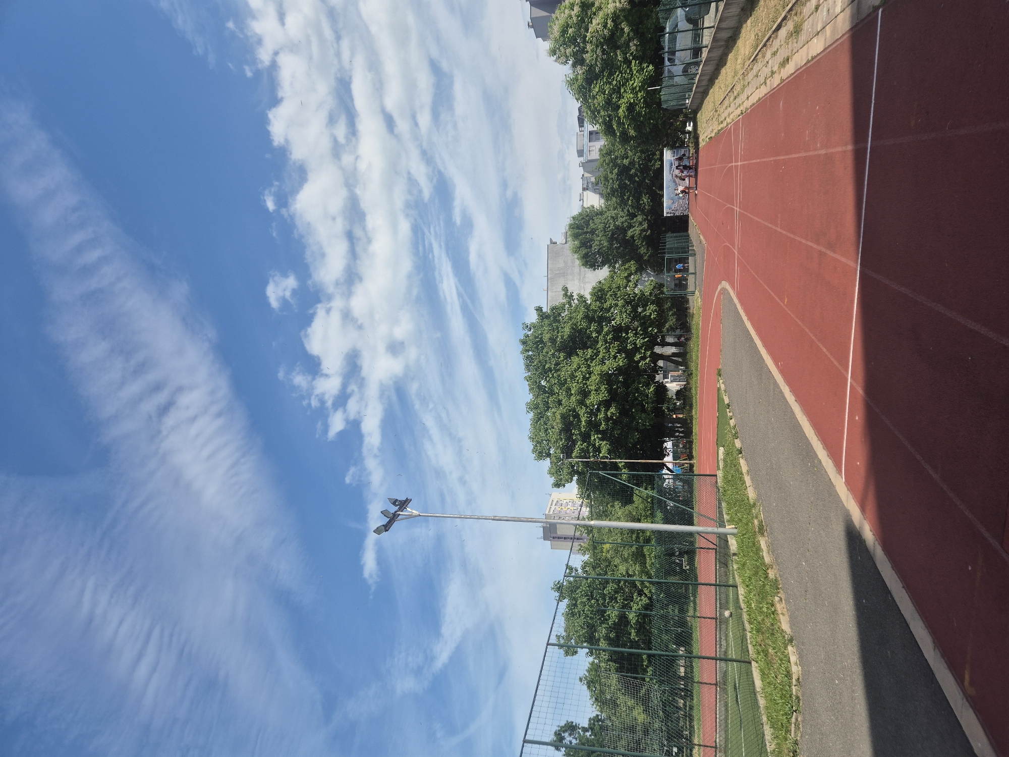 The image depicts an outdoor athletic track in an urban setting. The track is red with white lane markings. To the left of the track, there is a fenced-off area with green turf, possibly a sports field. A tall lamppost with multiple lights stands prominently on the left side of the frame. Behind the fence, a row of lush green trees borders the athletic facility. In the background, several multi-story buildings are visible, suggesting a city environment. The sky is bright blue with scattered white clouds. A group of people is gathered on the track in the middle ground, and a car is parked on the right side of the image. The overall scene suggests a sunny day at a public sports facility.