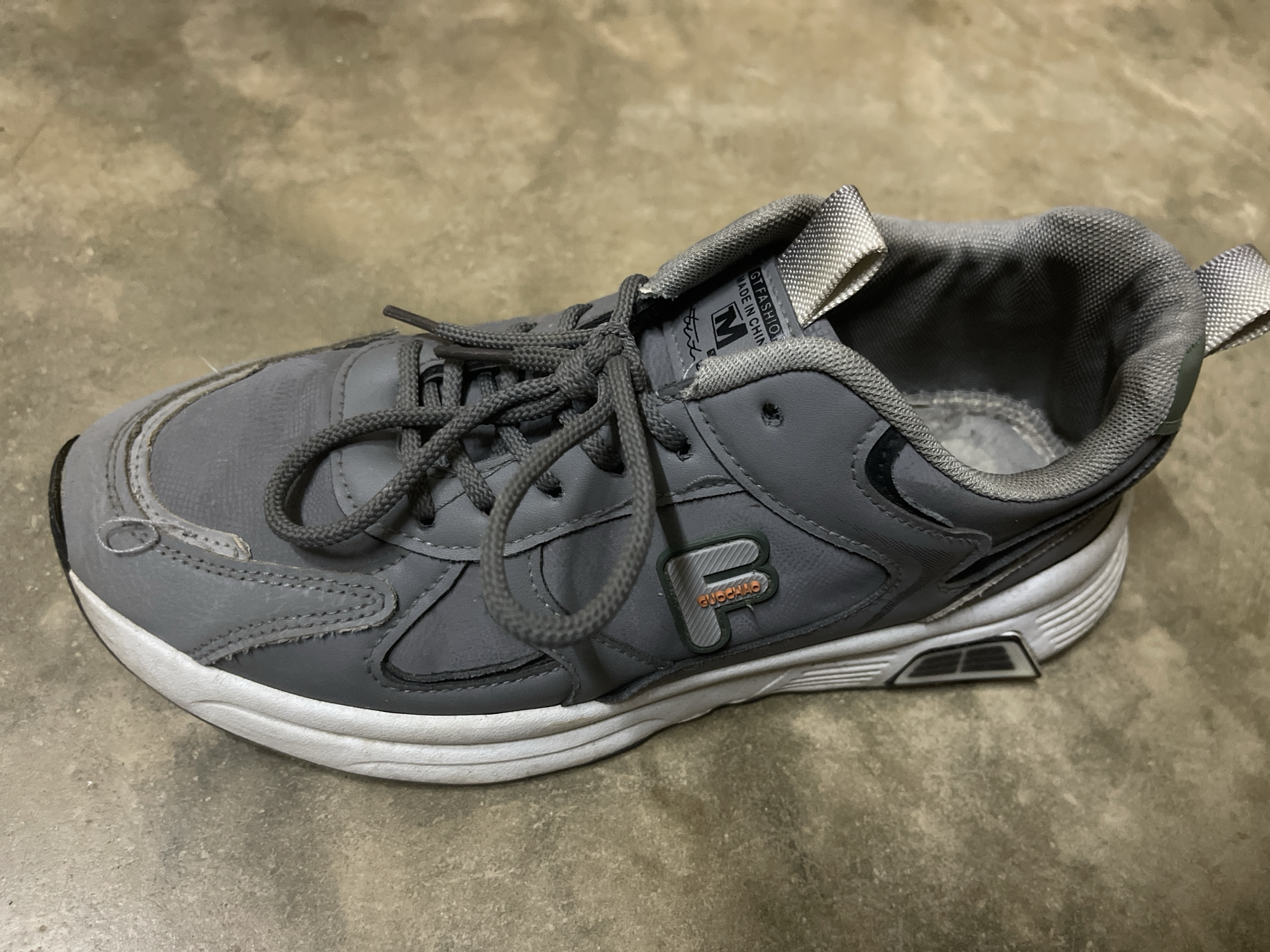 A single, well-worn gray athletic shoe, possibly a sneaker or walking shoe, is depicted resting on a textured light brown/gray concrete floor. No individuals are present, and no specific activity is taking place; the shoe is stationary. The setting appears to be an indoor or outdoor concrete surface, potentially in Dar es-Salaam, Tanzania, as per the provided context.
The shoe features gray laces, a white sole with integrated gray elements, and an upper constructed from what appears to be synthetic leather and mesh. Visible signs of wear are evident, particularly on the toe cap, the stitching along the side panels, and the slightly discolored white sole. The interior lining of the shoe shows wear and some fuzziness.
Notable details include text and logos on the shoe. On the tongue, a label reads "GT FASHION" and "MADE IN CHIN", likely indicating "MADE IN CHINA", alongside a distinct "M" logo within a square. On the side of the shoe, there is a stylized logo resembling an outlined 'F' in green with an orange-brown fill, and smaller text "GUOCHAO" underneath it. Silver-gray fabric loops are present on the front and back of the shoe's collar. The time of day and weather conditions are not discernible from the image.