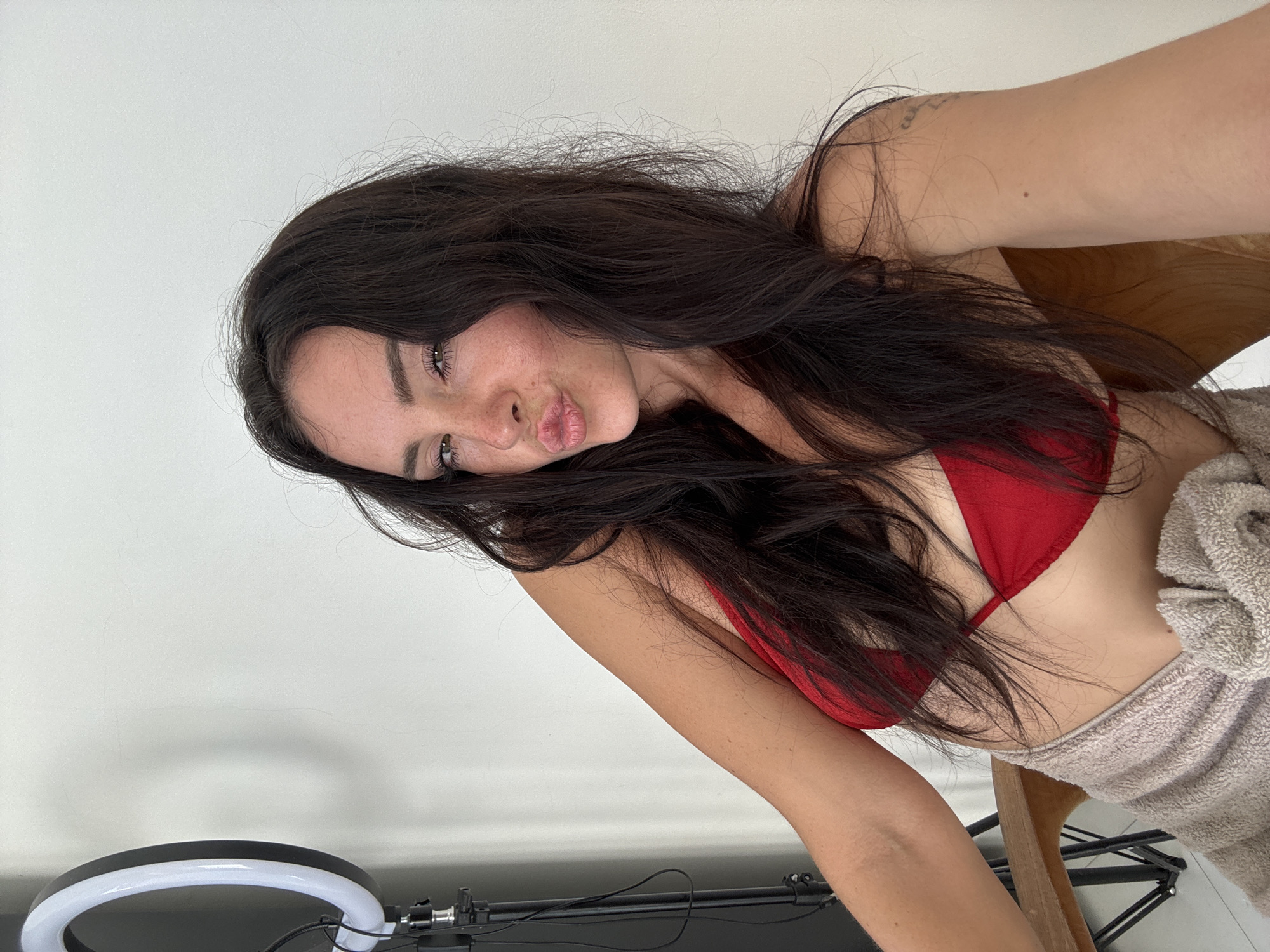 A young woman with long, dark hair, wearing a red bikini top and a beige towel draped around her waist, poses for a selfie. She is seated in a wooden chair against a plain white wall. To the left of the frame, a ring light on a tripod is visible. The woman has her right arm raised behind her head, and she is puckering her lips. Her expression is playful and engaging. A tattoo is visible on her right upper arm. The environment appears to be indoors, possibly a studio or a room in a house. The overall impression is one of casualness and self-expression.