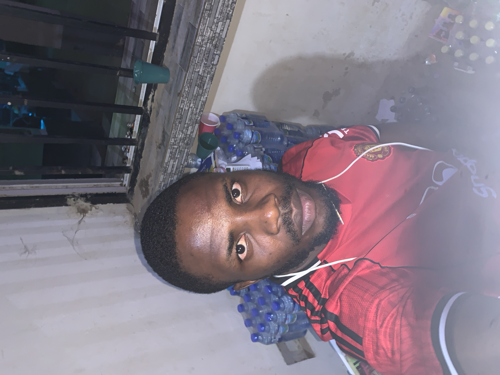 A close-up portrait of a dark-skinned man with short hair and a trimmed beard, wearing a red and black striped athletic shirt. He has earbuds in his ears. Behind him are stacks of plastic water bottles, a few plastic cups, and what appear to be packaged goods. The background also includes a window with a black metal frame and a section of a tiled wall. The location appears to be indoors, possibly in a storage area or back room based on the setting. While it is mentioned that the location is New Stadium, Nigeria, that cannot be forensically confirmed solely from this image.