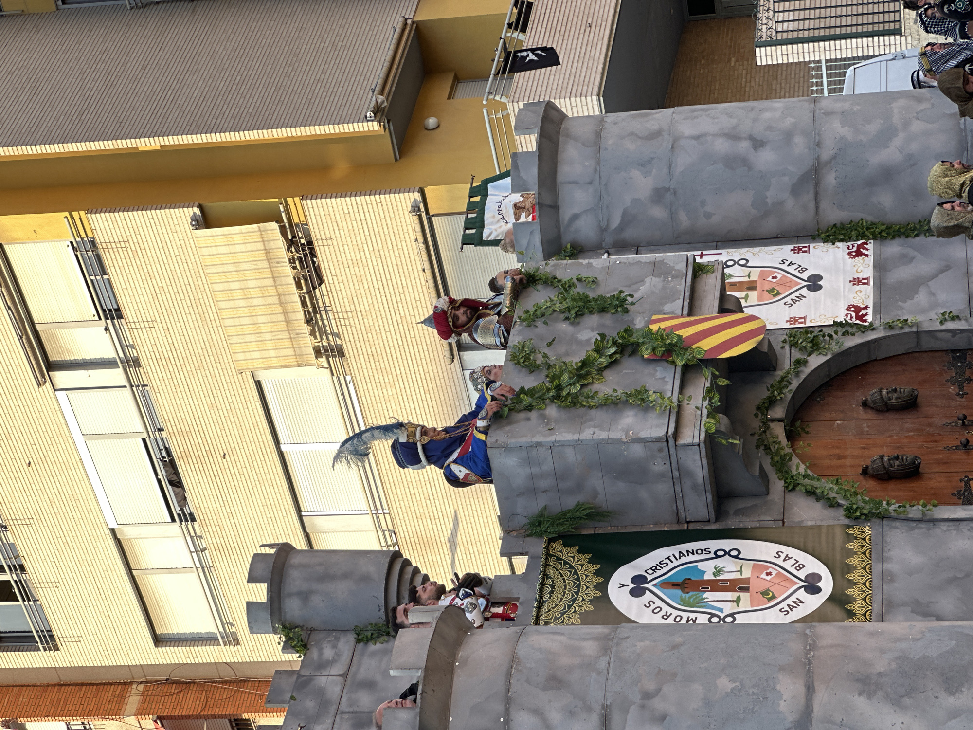 The image captures a scene from a historical reenactment or festival, likely the "Moros y Cristianos" (Moors and Christians) celebration, given the banners that read "MOROS Y CRISTIANOS SAN BLAS". The event appears to be taking place on a street in Alicante, Spain, as suggested by the architectural style of the building in the background, which features light-colored brickwork and modern windows with roller shutters, indicative of urban Spanish construction.
The foreground is dominated by a large, grey, castle-like structure, constructed with rough-textured panels. This structure has battlements and is adorned with artificial ivy. Two figures in elaborate historical costumes are visible on the castle's upper level. One figure on the right wears a red and gold turban and ornate armor, holding a shield. To his left, another figure sports a blue and gold headdress with a prominent feather and a blue and red tunic. They are both looking out over the scene.
Below them, on a lower level of the castle structure, a large banner with an oval crest featuring a lighthouse, palm trees, and flags is displayed. Another banner with a shield displaying red and yellow stripes is also visible. A large wooden door with carved faces is at the base of the structure.
In the background, a multi-story building with cream-colored brick and several windows is visible. Balconies with metal railings are present on the building. Several people, some in traditional headscarves, are visible at the very bottom of the image, suggesting a crowd or participants in the event. A white van is parked behind the castle structure, partially obscured. The overall atmosphere suggests a lively and colorful celebration.