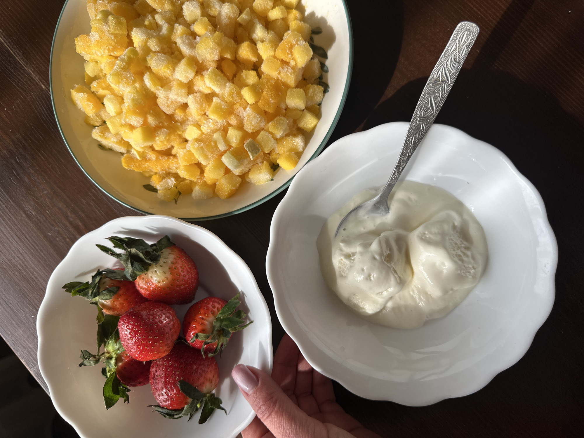 The image shows a table setting with three bowls containing different food items. The items are strawberries, diced mango, and a white creamy substance, likely yogurt or ice cream. A silver spoon rests in the bowl of the creamy substance. A hand is visible holding the bowl of strawberries. The scene is indoors, and the table appears to be dark wood. There is no indication of the city of Chaikovsky, Russia, or any specific location.