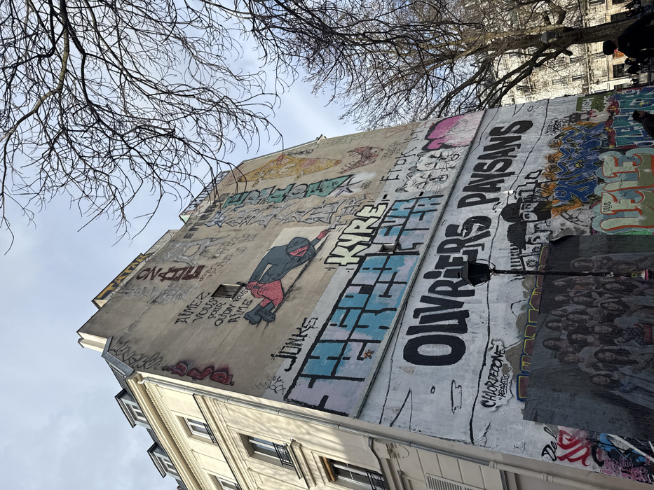 This image depicts a street scene in Paris, France, centered on a large building facade extensively covered in graffiti and murals. To the left, a multi-story Haussmannian-style building features a light-colored, ornate facade with numerous windows, dark frames, and light-toned shutters. To the right, an adjacent, tall, windowless concrete or plastered wall serves as a canvas for a dense collection of urban art.
The graffiti wall displays multiple layers of artwork. Towards the top, text reads "AIMEZ VOUS POUR QU'ON VOUS AIME" (Love yourself so that you are loved), below which a large graphic shows a figure in a red top and dark pants sliding diagonally downwards. Mid-wall, prominent block letters include "JONKS" and "KYRE," above the large black sans-serif text "OUVRIERS PAYSANS" (Workers Peasants). Below this, a black, ornate street lamp stands in the foreground, partially obscuring the lower murals. To the left of the lamp, a rectangular mural features a group of approximately 20-25 young women, primarily with long hair and light-colored attire, arranged in rows. To the right of the lamp, additional graffiti includes stylized faces, a bearded figure with a cap, and various blue, orange, and black tags.
Bare, deciduous tree branches with no leaves frame the upper right portion of the image, silhouetted against an overcast or partly cloudy sky. In the far lower right, several indistinct human figures are visible on a sidewalk or street, with another classic Parisian building faintly visible in the background.