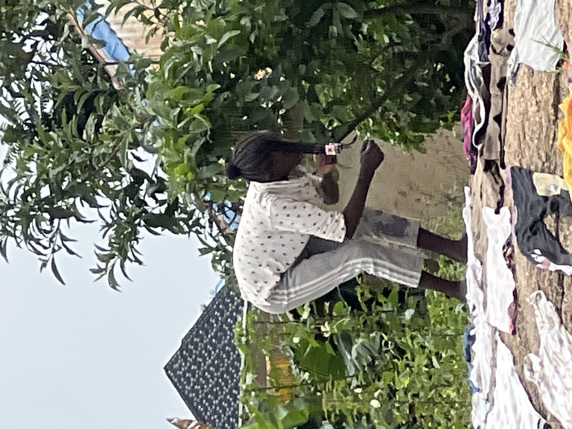 In the city of Dong, Nigeria, a young person with braided hair tied in a bun, wearing a white shirt with a pink polka dot pattern and gray athletic pants with white stripes, is crouched down. They are looking at and holding a mobile phone with their right hand, which is extended towards a tree. Their left hand is also outstretched. The person is standing on a surface covered with assorted laundry, including white t-shirts, striped towels, and other garments. In the background, lush green foliage dominates the scene, with glimpses of buildings and a dark, textured roof. The sky above is overcast. The overall impression is of an outdoor domestic activity.