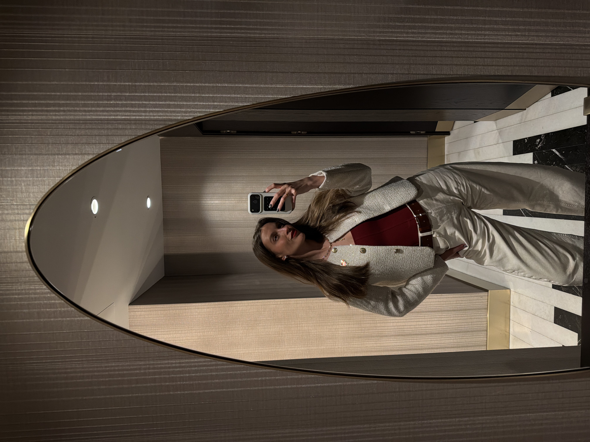 A woman is reflected in an oval mirror. She is holding a smartphone and is wearing a maroon shirt, a cream-colored blazer with gold buttons, cream-colored pants with a brown belt. The background shows wallpaper and a black and white tiled floor. The location is most likely an interior space, possibly in Budva, Montenegro.