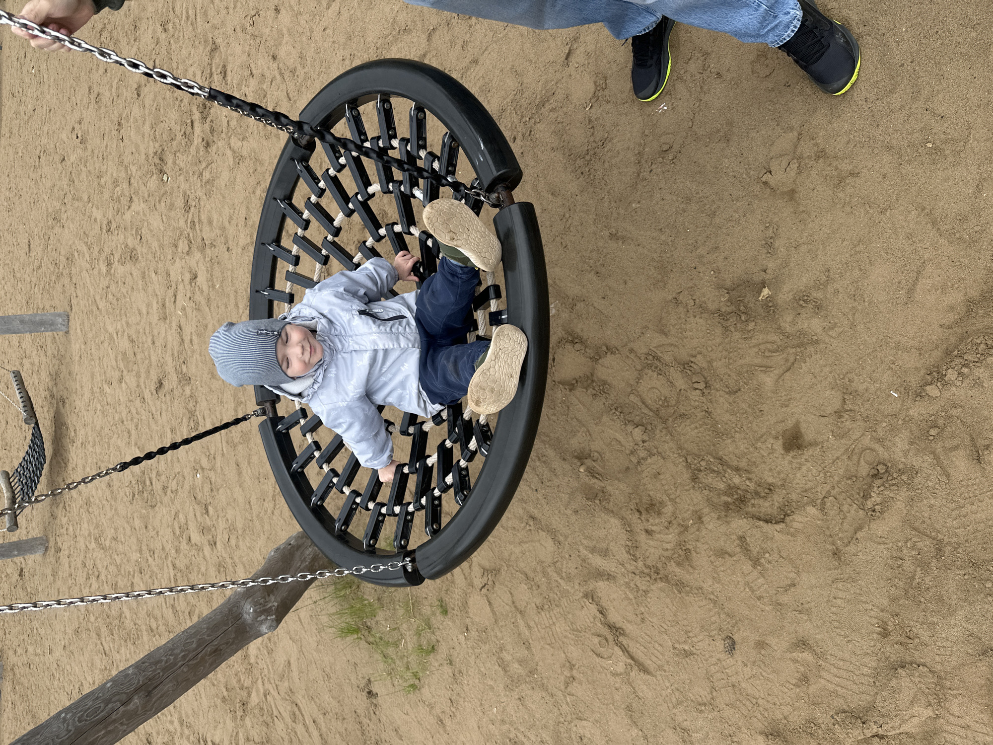 The image displays an outdoor playground scene, reported to be in the city of Chaikovsky, Russia. A young child is positioned centrally, seated within a black-framed saucer swing that features a woven net base of dark straps and lighter ropes. The child wears a light grey hooded jacket, dark blue trousers, and a grey and white striped knit beanie. Light-colored, possibly beige or light green, shoes with textured soles are visible on the child's feet, resting on the swing's netting. The child's head is tilted slightly down and to the right, with a soft, content expression.
The saucer swing is suspended by two metallic chains. In the upper right section, an adult's hand is visible grasping one of these chains, indicating interaction with the swing. The adult's lower legs and feet are present in the bottom right corner, clad in blue jeans and black athletic shoes with distinct yellow-green soles.
A thick, weathered wooden support post is visible on the left side of the frame, to which one of the swing chains appears to be attached. In the background, another playground structure, consisting of a net hammock or bridge suspended between additional wooden posts, is partially visible. The ground is composed of light brown, finely textured sand, marked with various footprints and surface disturbances. Small areas of green grass grow near the base of the wooden post on the left. The lighting is diffused, consistent with an overcast sky.