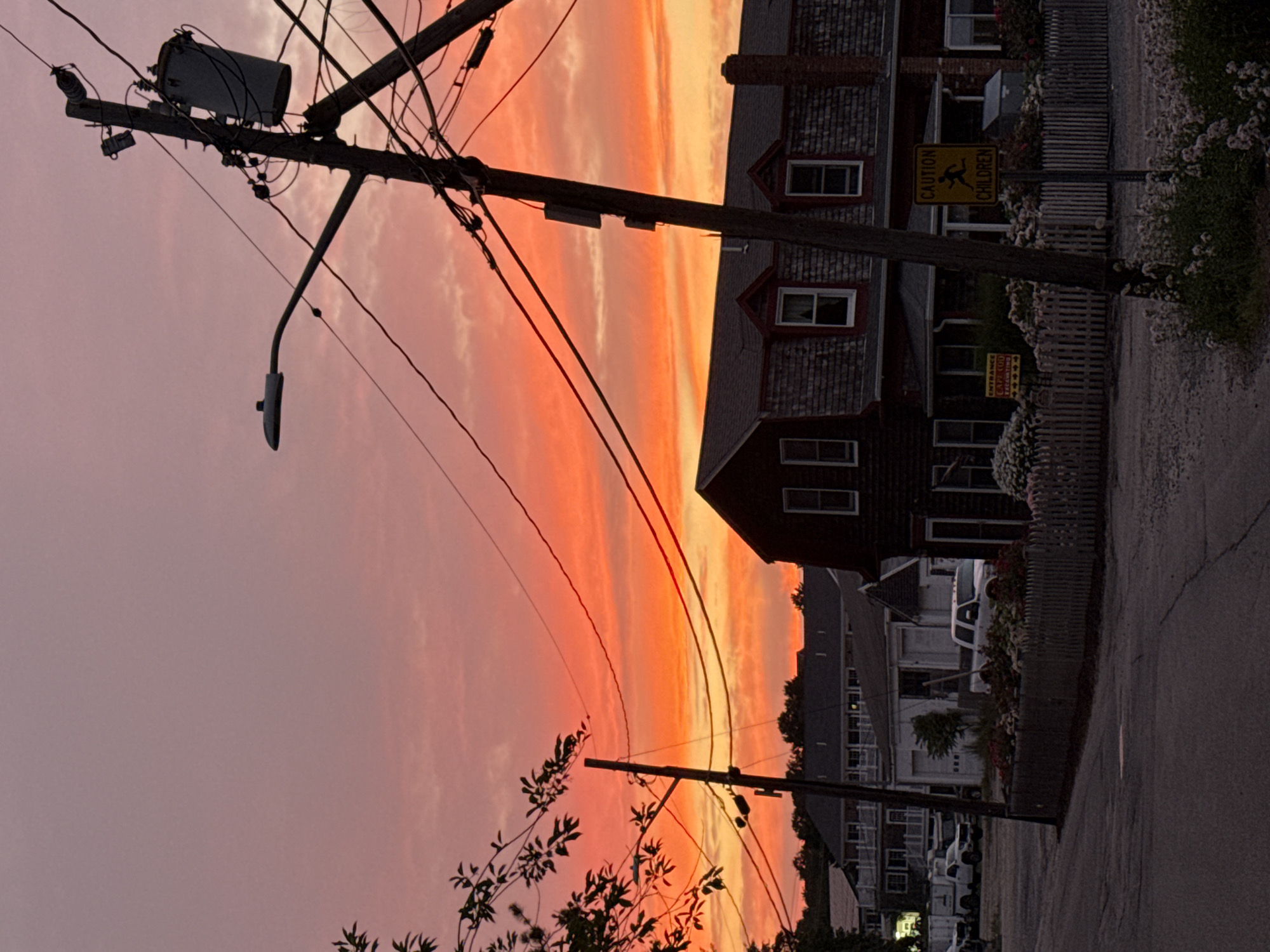 A street scene in Provincetown, Massachusetts, USA, is depicted at dusk. The sky is a vibrant gradient of orange and pink, with wispy clouds. In the foreground, a paved road leads into the scene, bordered by a low wooden fence and blooming white flowers. A utility pole with numerous wires and a street lamp dominates the right side of the frame. Behind the fence, several buildings are visible. To the right, a dark-colored, multi-story building with shingled siding and dormer windows stands prominently. A yellow caution sign indicating "CAUTION CHILDREN" is mounted on a pole in front of this building. To the left, a lighter-colored building with a garage door and a white truck parked in front is visible. Further in the background, more buildings and trees are silhouetted against the colorful sky. The overall atmosphere is tranquil, with the warm hues of the sunset creating a serene backdrop for the residential and commercial structures.
