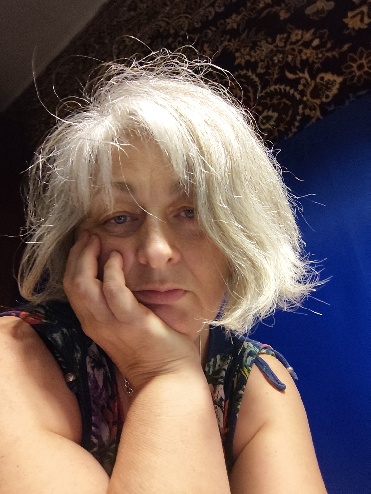 A woman with light-colored hair rests her chin on her hand, looking towards the camera. She is wearing a dark-colored top with a floral pattern. The background consists of a dark patterned fabric and a bright blue surface. The image appears to be a selfie taken indoors. There is no information in the image to suggest it is located in Krasnodar, Russia.