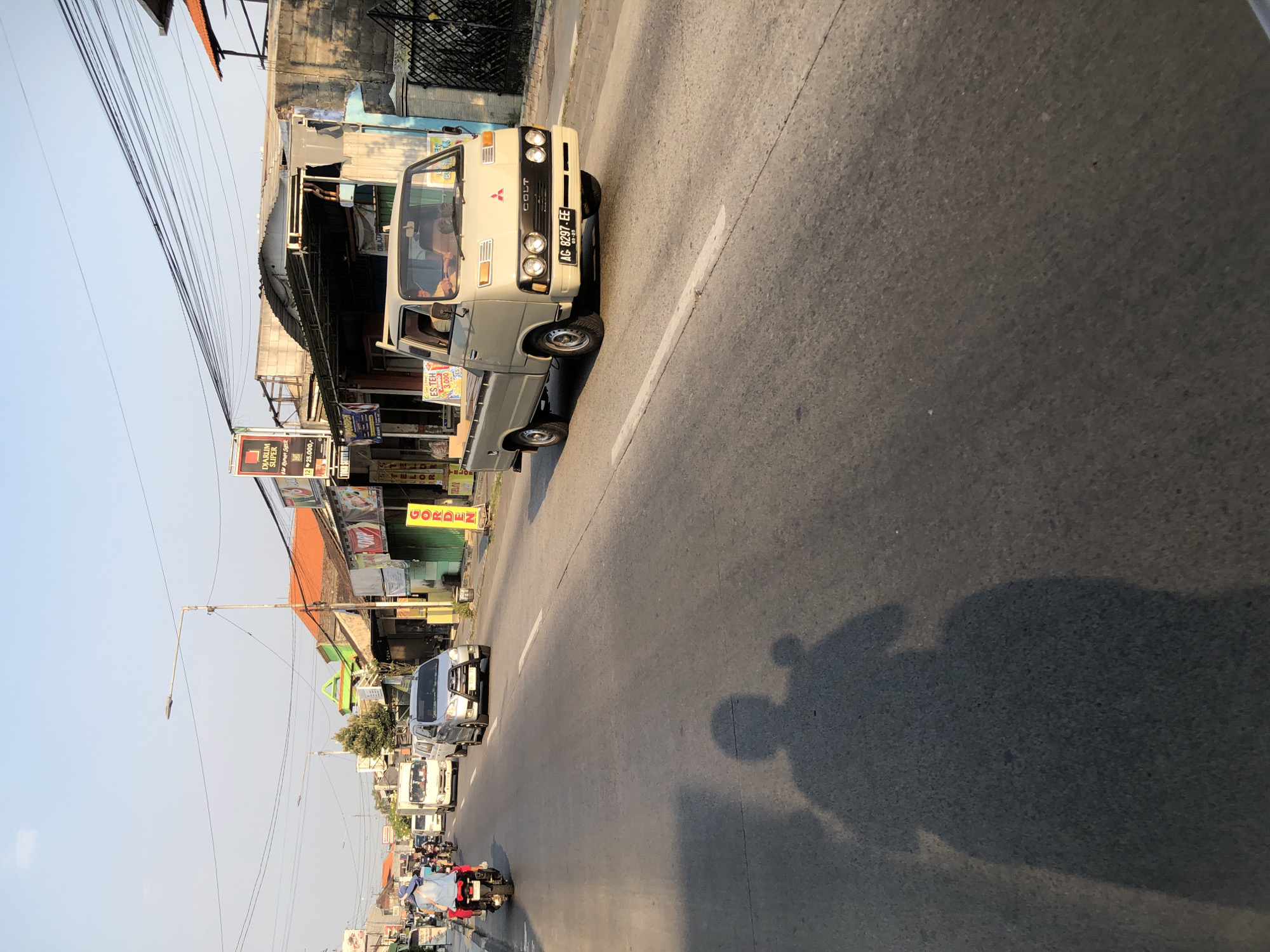 The image depicts a street scene in Boro, Indonesia, during daylight with a clear sky. In the foreground, a shadow of a person on a motorcycle is cast across the asphalt road. The road is populated with various vehicles, including a beige pickup truck with the brand "COLT" visible on its grille and license plate "AG 8297 EE," a silver SUV, several motorcycles, and a white van. On the right side of the road, buildings with shops are visible, featuring signs such as "GORDEN" and advertisements for "ES TEH." The buildings have tiled roofs and some exhibit exposed brickwork. Overhead, electrical wires crisscross the sky. The lighting suggests it is late afternoon, with the sun casting long shadows.