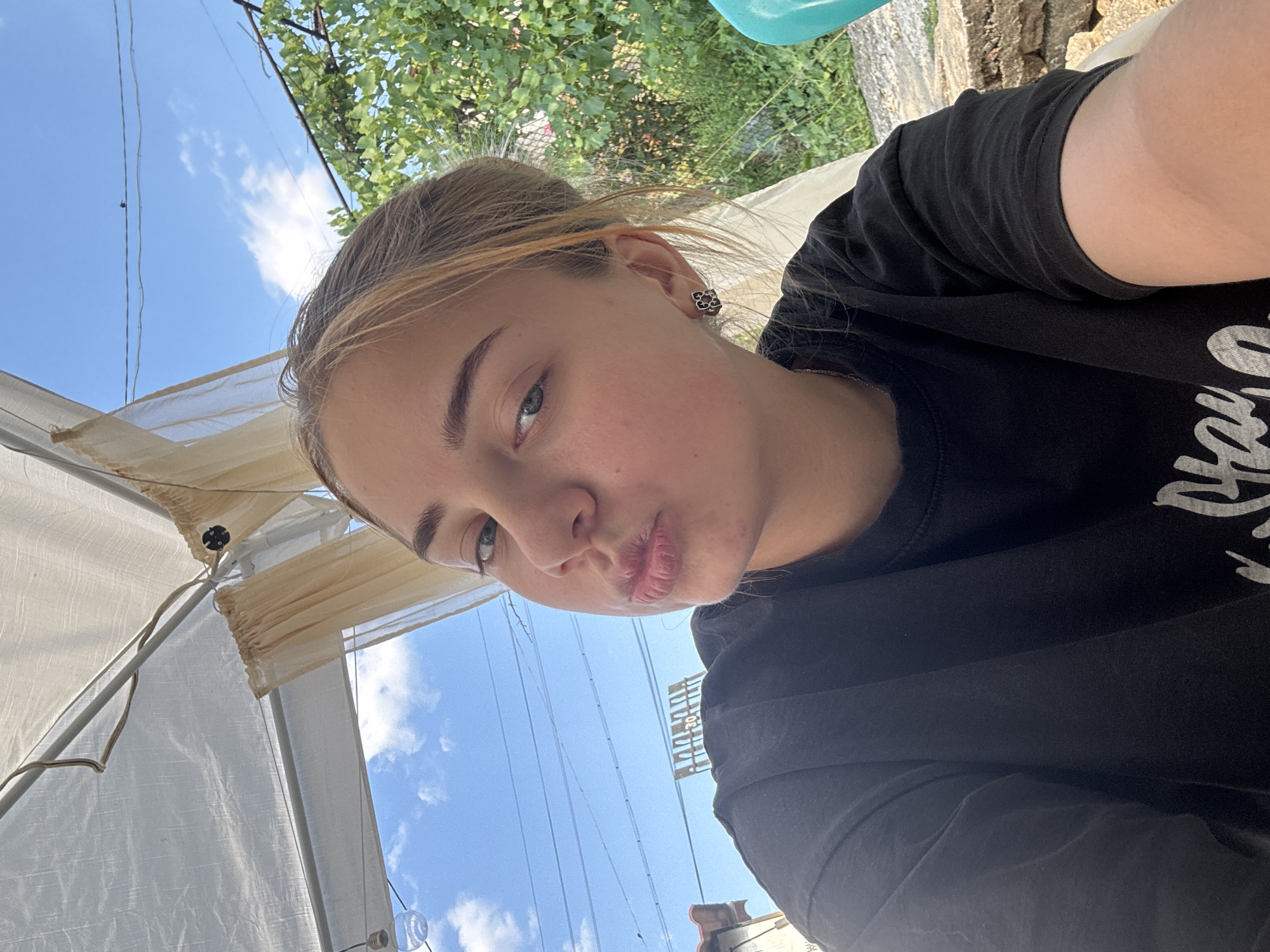 A young Caucasian female with blonde hair pulled back is taking a selfie outdoors under a light-colored canopy. She is wearing a black t-shirt and a black earring with a floral design in her right ear. Her lips are pursed for a kiss. In the background, there is a blue sky with white clouds, green foliage, and some overhead wires. A partial view of a building and some stone work is also visible. The image appears to have been taken during the day in a residential area of Sredets, Bulgaria.