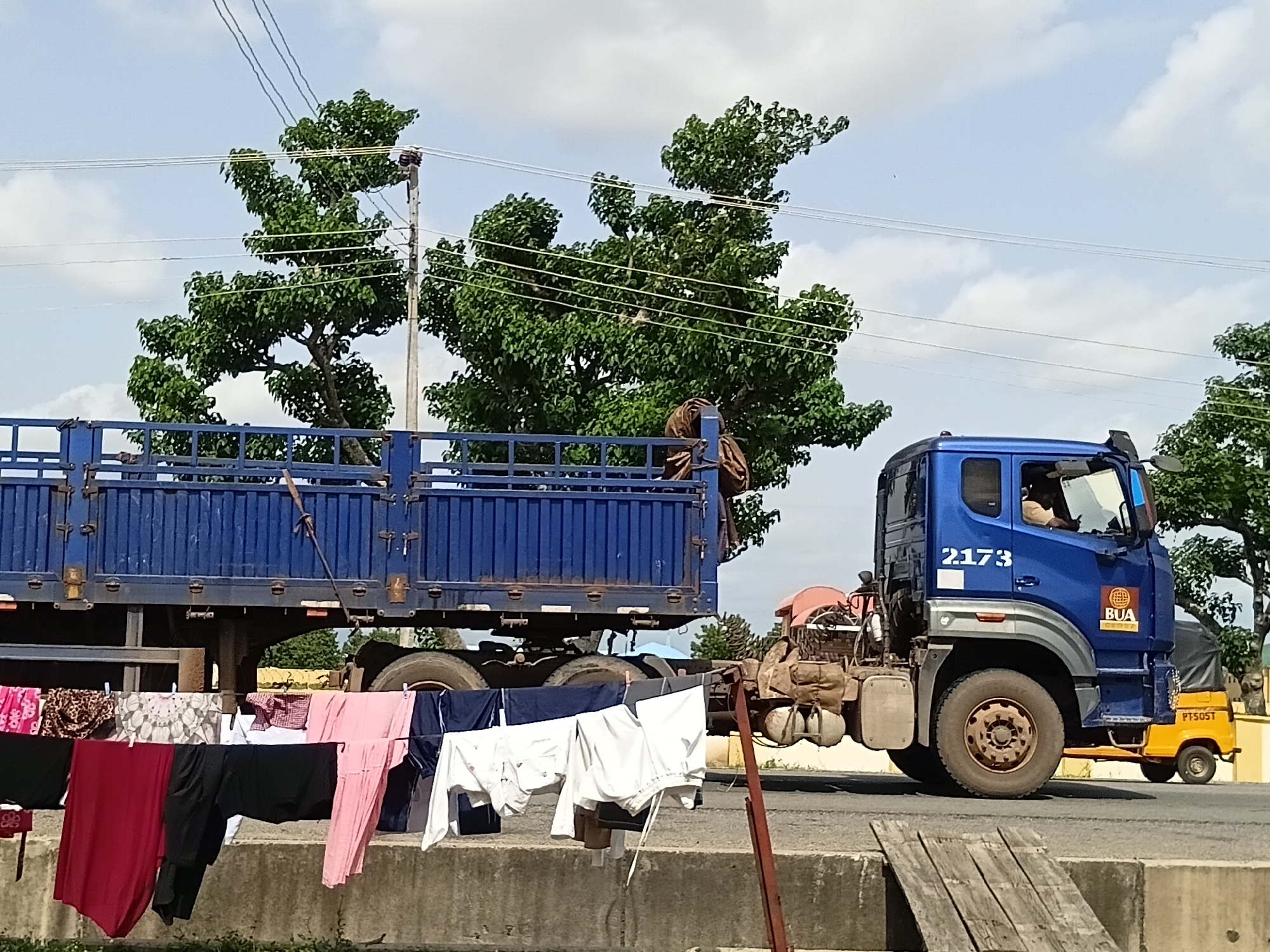 The image captures a scene on a paved road in Jalingo, Nigeria, under a partly cloudy sky. In the foreground, laundry is hung on a clothesline, with a variety of garments including a red skirt, black pants, a pink dress, and white shirts. A concrete barrier runs along the side of the road, with some wooden planks placed on it.
Dominating the midground is a large blue truck, its trailer filled with what appears to be bundled cargo covered by a brown tarp. To the right of the trailer, a smaller blue truck cab is visible, with the number "2173" displayed on its side and a "BUA" logo. A person is seated in the driver's seat of the blue truck cab. To the right of the blue truck, a small yellow three-wheeled vehicle, possibly a Keke Napep, is also present.
The background features lush green trees, some of which are quite large and have a distinct branching pattern. Electrical wires and a utility pole are visible against the sky. The overall impression is of a typical roadside scene in a Nigerian city, with commercial vehicles and domestic activities coexisting.