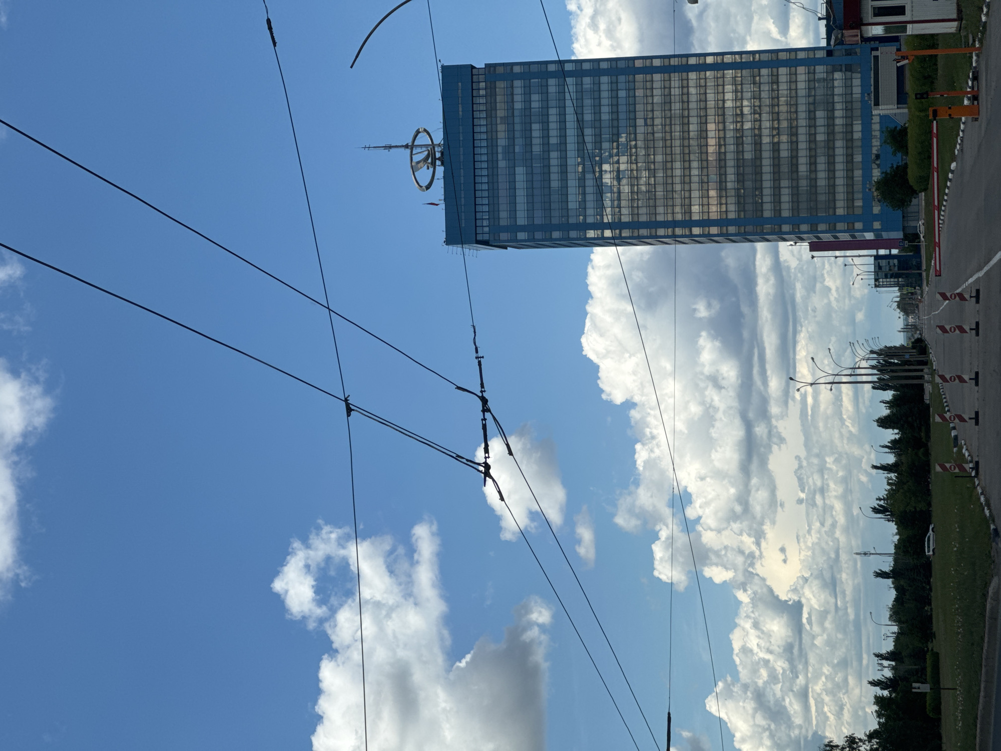 The image shows a tall building with a ring-shaped structure on top. Overhead power lines intersect the view, set against a backdrop of a blue sky with cumulus clouds. A road with traffic cones lines the right edge of the frame. Trees and streetlights are visible along the road.