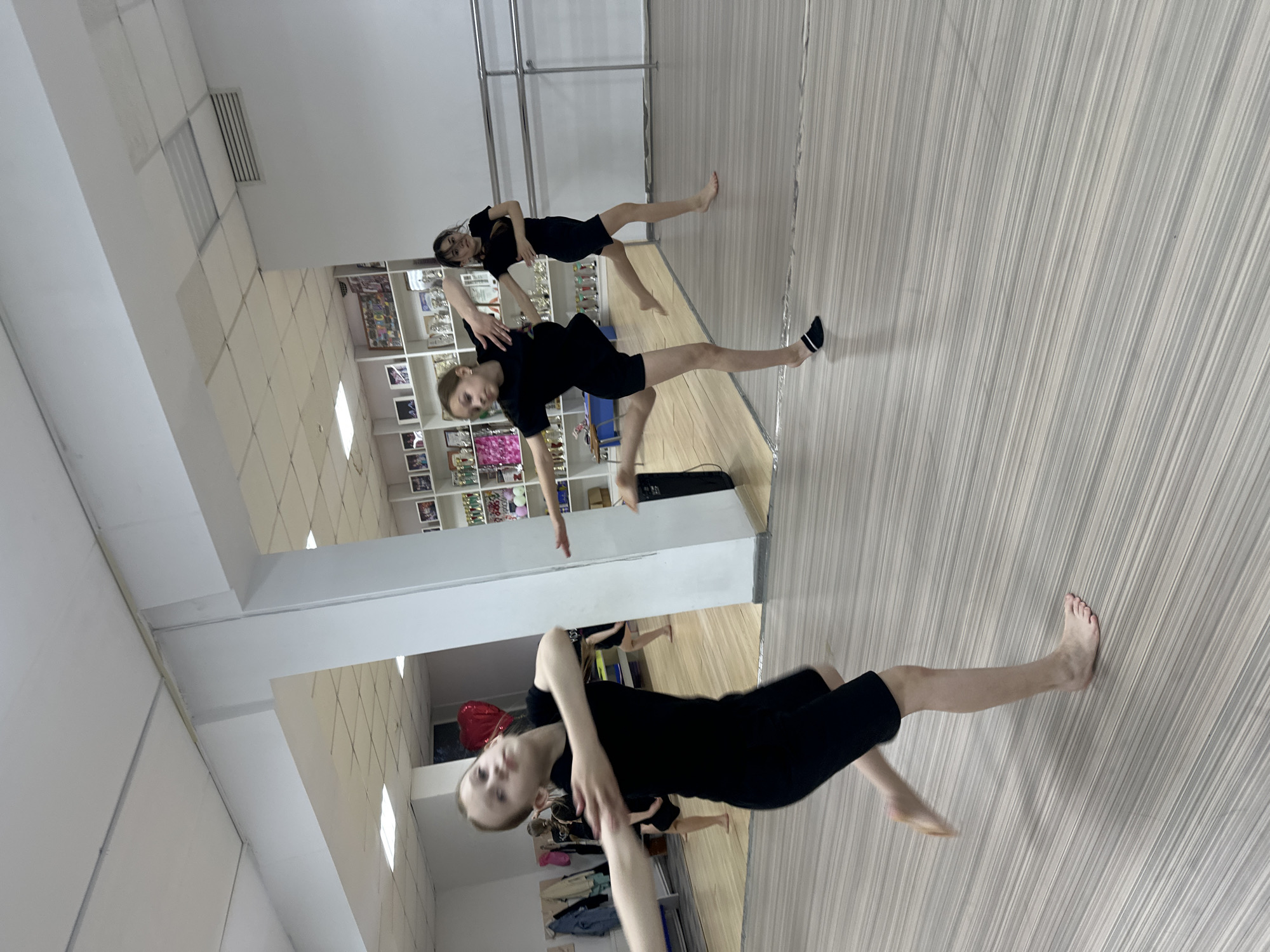 A dance studio in Votkinsk, Russia. Three young women are practicing a dance routine. They are dressed in black clothing. The studio has a wooden floor, mirrors, and a ballet barre. A shelf displays trophies and photographs.