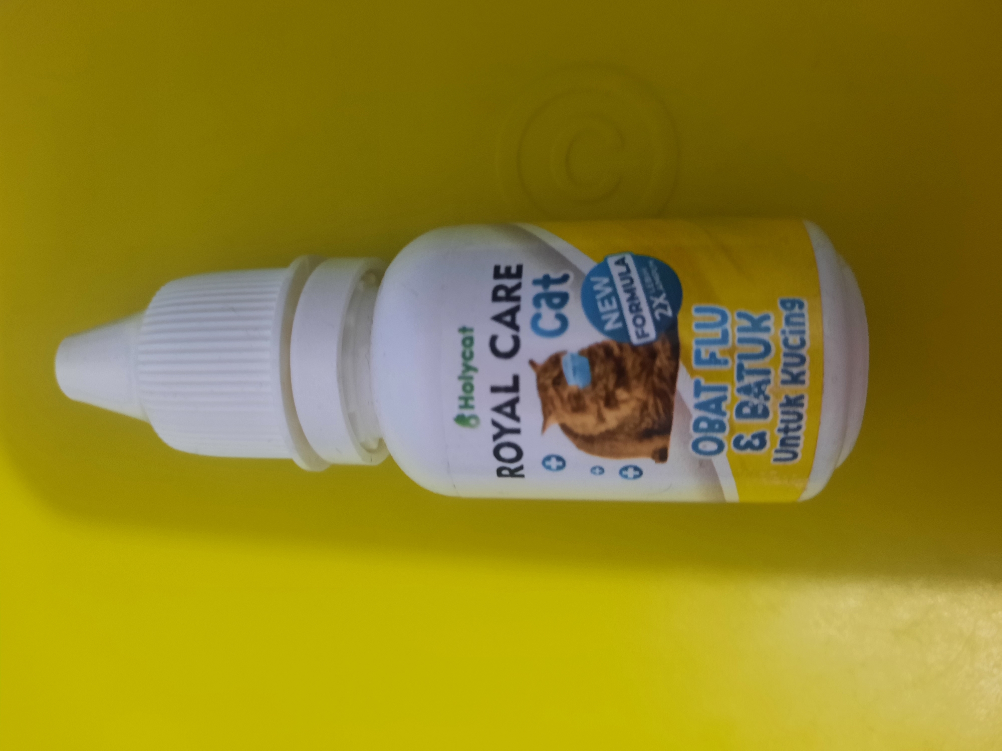 The image shows a close-up, top-down view of a small white bottle with a dropper top, resting on a bright yellow surface. The bottle is predominantly white with a yellow label wrapping around its body. The label features text and an illustration. The text on the label reads "Holycat" in a small green font, followed by "ROYAL CARE" in large, bold black letters. Below this, in smaller blue text, is "Cat". To the left of the "Cat" text is a graphic of a brown cat wearing a light blue mask. Below the cat illustration and the text "Cat" is a blue circle with "NEW FORMULA" in white text and "2X" with smaller text below it that is difficult to read. The bottom of the yellow label contains text in blue and white, reading "OBAT FLU & BATUK Untuk Kucing", which translates to "Flu & Cough Medicine For Cats". The yellow surface has a subtle circular indentation or shadow in the background to the right of the bottle. The lighting appears to be from above and slightly to the left, casting a shadow of the bottle onto the yellow surface. There are no people or animals visible in the image, and the scene is indoors, likely a tabletop or similar flat surface. There is no information in the image that could be used to determine its location in Lhokseumawe, Indonesia.