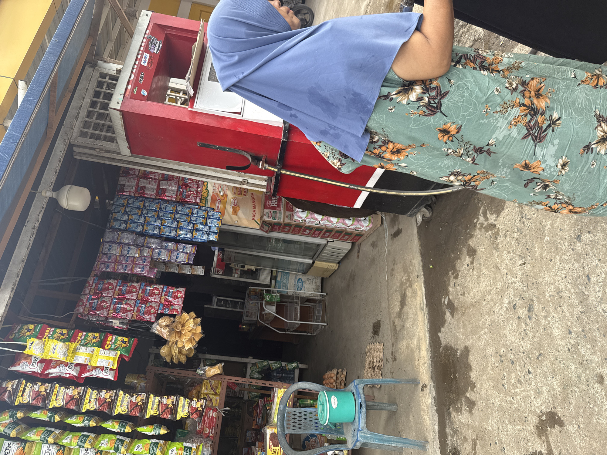 This image captures a street-level view of a small convenience store in Ajuen, Indonesia. In the foreground, a person wearing a blue hijab and a green floral dress stands near a red gasoline pump. The store's facade is lined with shelves displaying a variety of packaged goods, including snacks, drinks, and household items. Several strings of brightly colored snacks hang from the ceiling. A blue plastic chair with a green bucket on it is placed on the concrete ground, with a stack of egg cartons nearby. The ground appears damp and slightly wet in places. The overall scene depicts a typical day at a local Indonesian market.