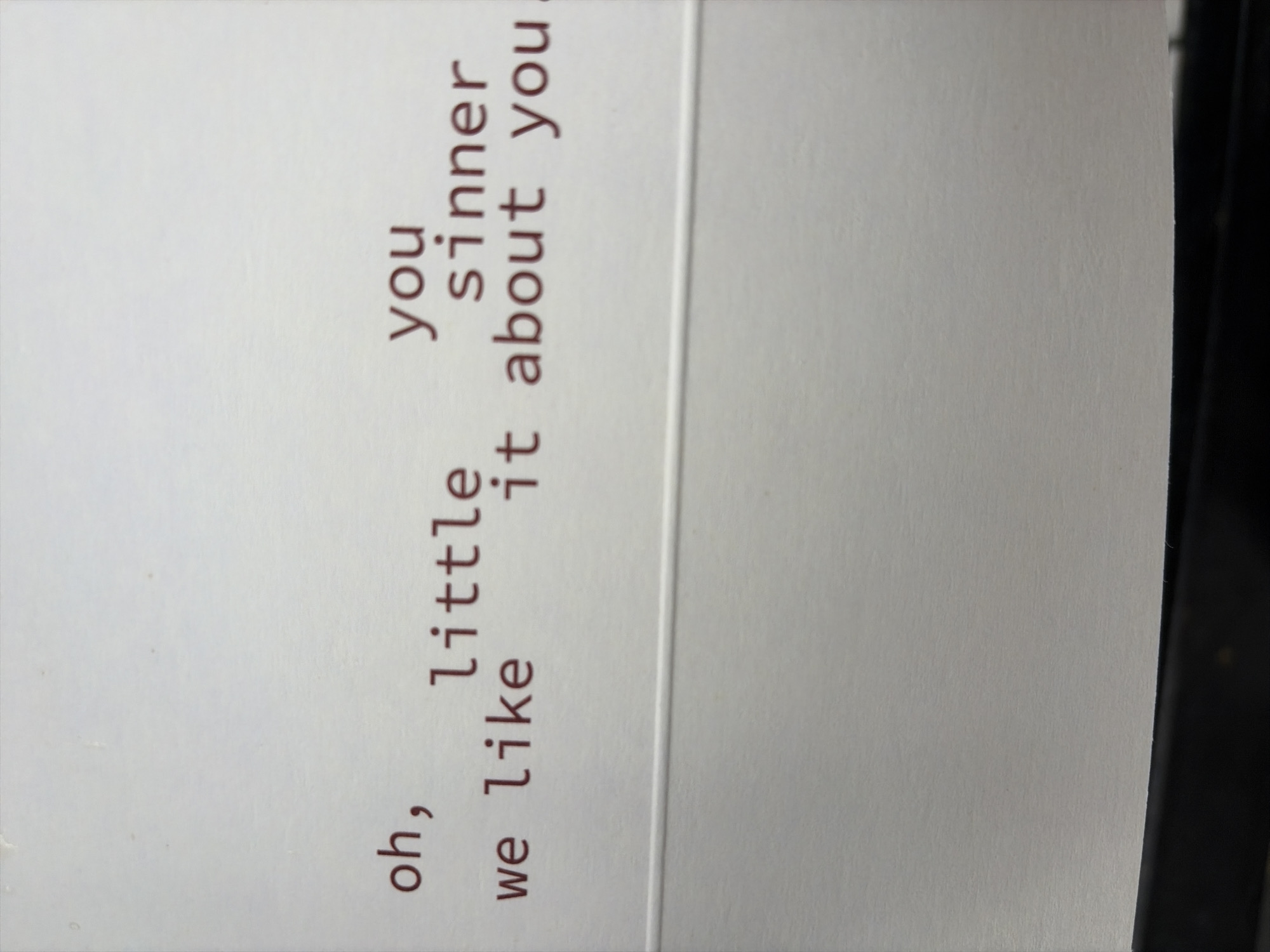 The image is a close-up, eye-level shot of white paper with red typewriter text. The text is split into two lines. The top line reads "you sinner". The bottom line reads "oh, little we like it about you". There is a horizontal crease across the paper below the text. The background is a plain, textured white. The bottom of the image shows a black, curved object, likely the platen of a typewriter. The location of Oxentea, Moldova is not visible or inferable from the image.