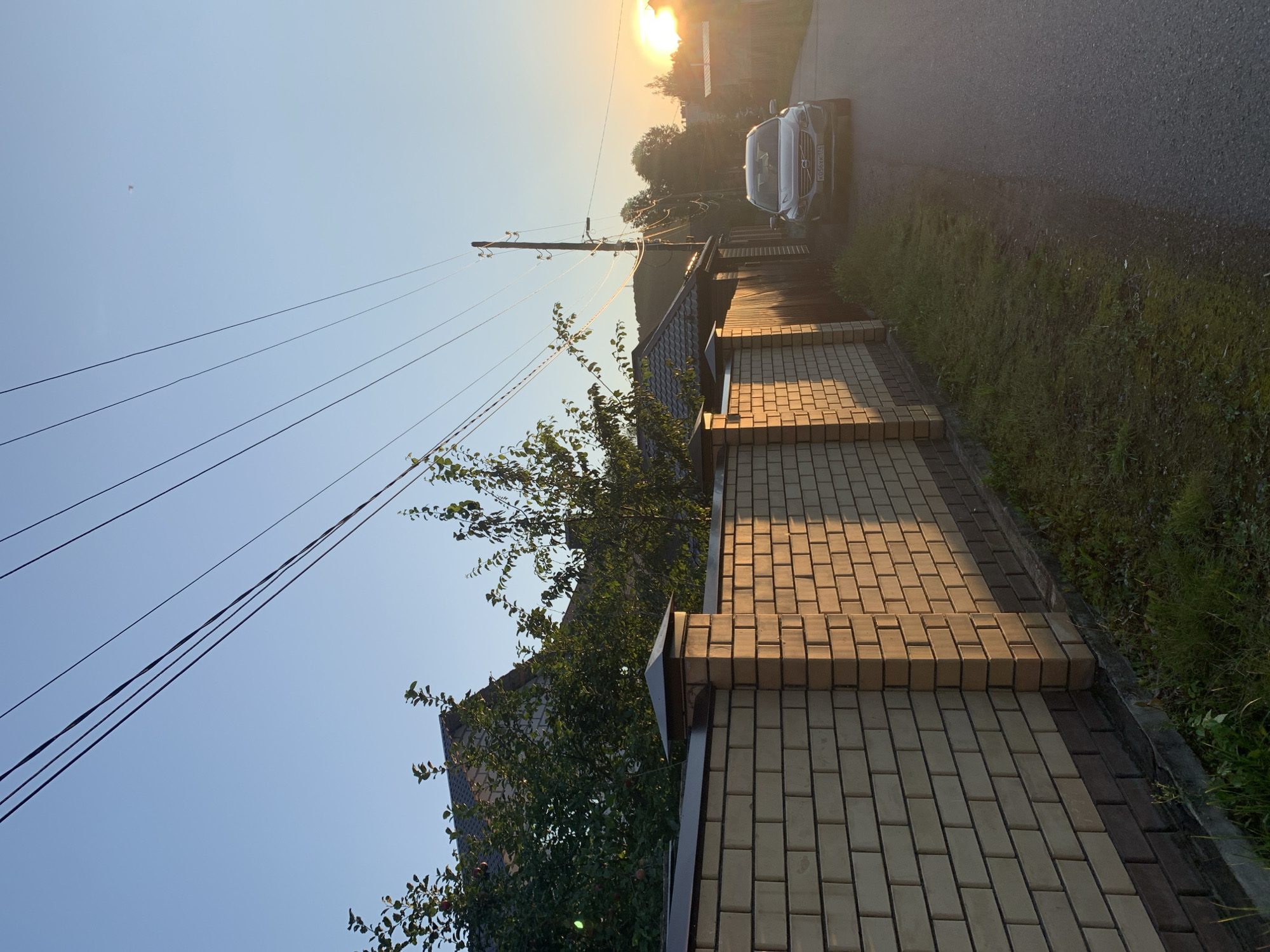 This is a daylight, eye-level, medium shot of a residential street in Asha, Russia, during sunset. A light-colored brick fence dominates the left side of the frame, casting long shadows from the setting sun. Behind the fence, a dark-roofed house and lush green trees are visible. A gray Volvo SUV is parked on the asphalt road on the right side of the image, facing the viewer. The sky is clear and blue, with a gradient of orange and yellow near the horizon where the sun is setting. Power lines crisscross the sky, and a utility pole stands on the right side of the road. The road appears to be a quiet, residential street with sparse vegetation.
