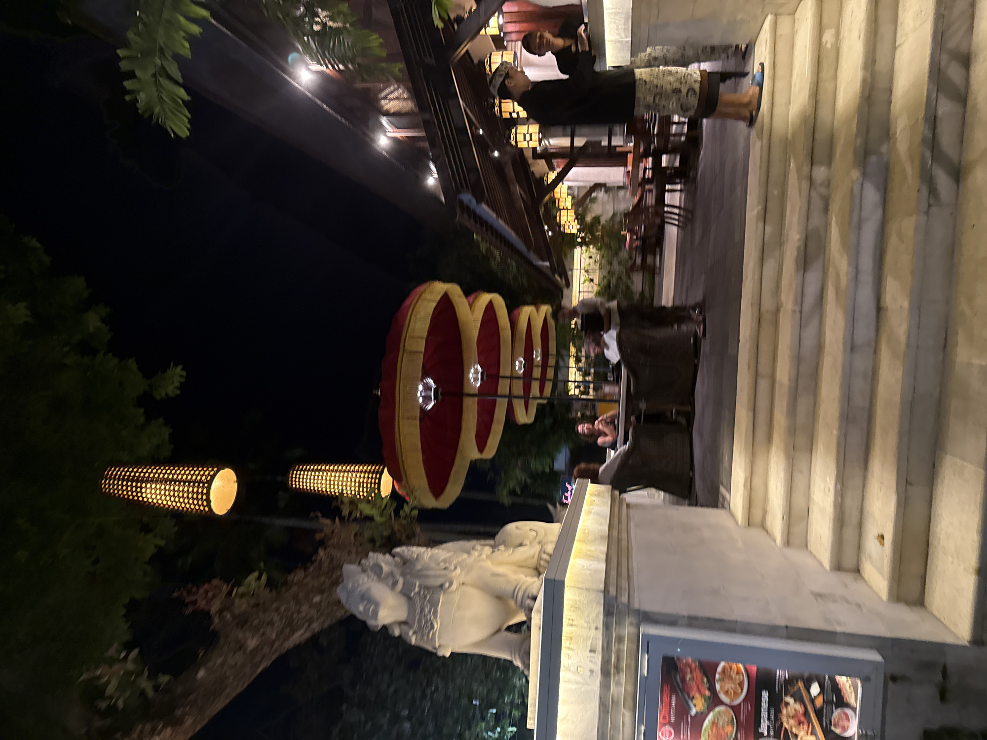 The image captures an evening scene at a restaurant in Denpasar, Indonesia, characterized by a blend of Balinese architectural elements and outdoor dining. In the foreground, a set of wide stone stairs leads up to the restaurant's entrance. To the left of the stairs, a illuminated menu board displays images of Chinese and Japanese cuisine. A large, white stone statue resembling a mythical creature, possibly a lion or guardian, sits atop a raised platform to the left of the stairs, illuminated by soft lighting.
The dining area is visible behind the statue, with tables and chairs arranged on a paved patio. Several patrons are seated at the tables, engaged in conversation. Overhead, a series of decorative, cylindrical lamps hang from poles, casting a warm, amber glow. Further back, a building with a traditional Balinese roof structure houses the restaurant's interior, with more lanterns visible. Red and yellow layered umbrellas are positioned above some of the outdoor seating areas, adding a touch of cultural flair.
Two men, appearing to be staff members, are near a counter or bar area. One is wearing a traditional patterned sarong and a dark shirt, while the other is dressed in a dark shirt and checkered pants. The lighting is dim, indicating it is nighttime, with artificial lights illuminating the scene. The overall atmosphere suggests a lively and welcoming dining establishment.