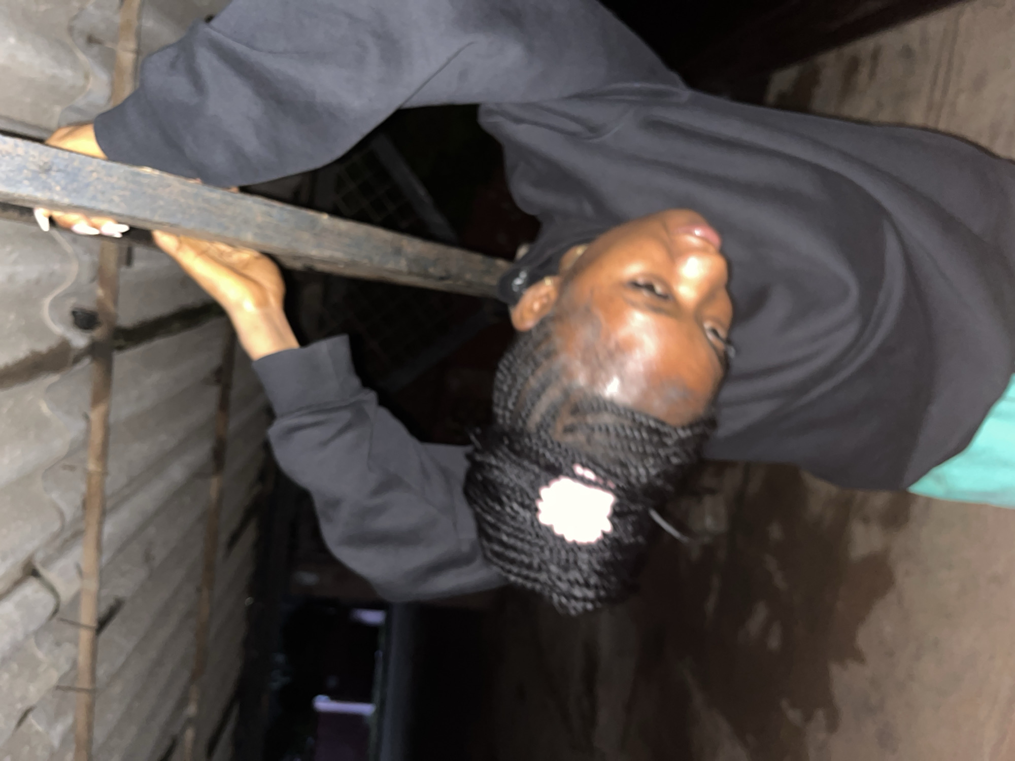 The image captures a close-up, low-angle shot of a young Black person with braided hair, adorned with a light-colored scrunchie, leaning back and holding onto a vertical metal pole. The person is wearing a black hoodie and teal shorts. Their head is tilted downwards, and their gaze is directed away from the camera. The background reveals a corrugated metal roof structure above, supported by wooden beams. Below the roof, a dark, unpaved ground surface is visible, with what appears to be patches of water or dampness. The overall lighting suggests an outdoor, possibly nighttime, setting. The scene is set under a rudimentary shelter, characteristic of some urban or peri-urban environments. This type of structure, often seen in less developed areas, can be found in various parts of Jos, Nigeria, particularly in informal settlements or areas with limited infrastructure, providing basic protection from the elements.