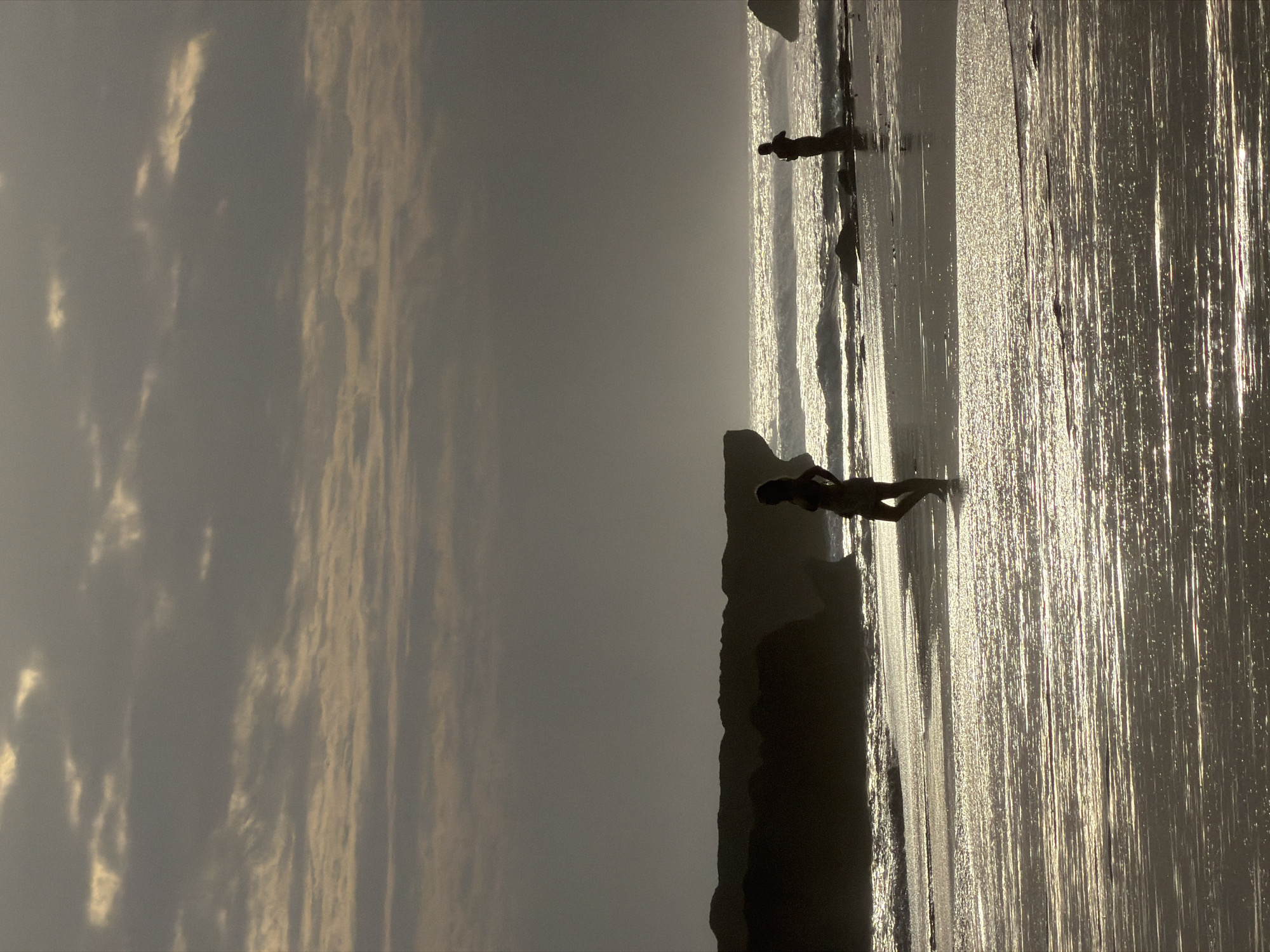 The image depicts a low-angle, silhouetted view of a beach in North Kuta, Indonesia, likely at sunset or sunrise due to the dramatic lighting. Two individuals are visible, both appearing as dark silhouettes against the bright horizon. In the foreground, to the left, a person stands near the water's edge, facing the ocean. Further back, closer to the center of the frame, another person is positioned on the wet sand. The ocean is visible in the mid-ground, with breaking waves creating a shimmering effect. Dark, rocky formations line the shore to the left, partially obscuring the horizon. The sky is filled with muted, cloudy formations, with some lighter patches suggesting the direction of the sun. The wet sand in the foreground reflects the light from the sky, creating streaks of shimmer.