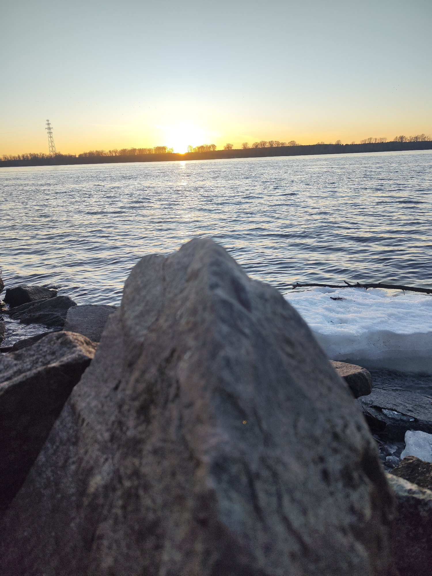 A landscape photograph depicts a body of water at sunset. The foreground features a cluster of rocks, one of which is prominently out of focus. The body of water is rippled, reflecting the sunset. In the background, a strip of land with bare trees is visible against the setting sun. An electrical transmission tower is visible to the left. A small patch of ice sits on the shoreline. The location is Noviy, Russia.