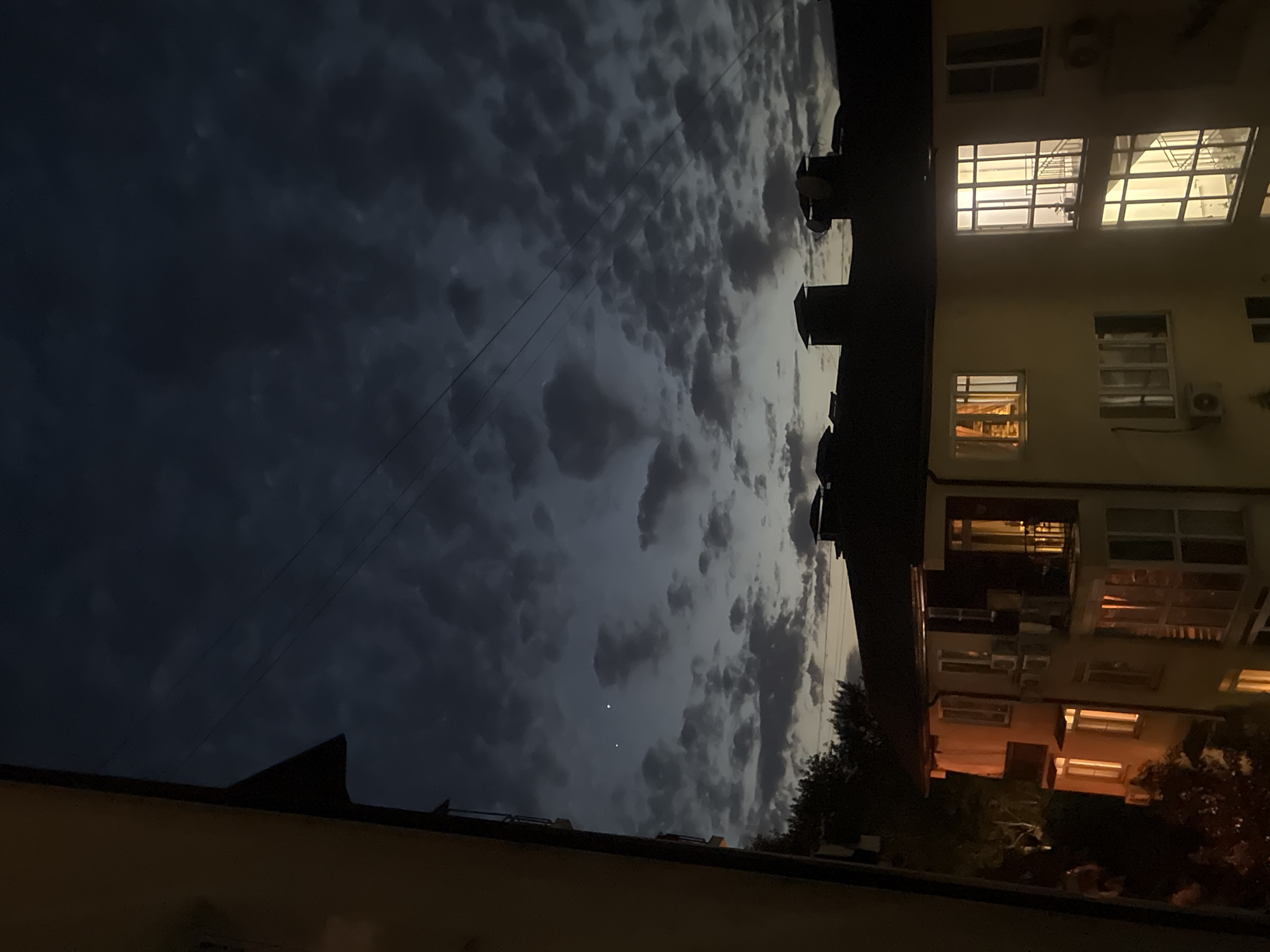 The image depicts a twilight scene over a multi-story building, likely an apartment complex, in Sochi, Russia. The sky is a deep blue, partially obscured by scattered, dark clouds. Two bright celestial bodies, possibly planets or bright stars, are visible in the upper left quadrant of the sky. Power lines crisscross the sky above the building. The building itself is illuminated from within, with several windows displaying warm, yellow light, suggesting that residents are home. The architecture appears to be modern, with balconies and multiple window units visible. The lower part of the image shows dark trees, possibly part of a park or residential landscaping. The overall impression is one of a quiet evening in an urban or suburban setting.